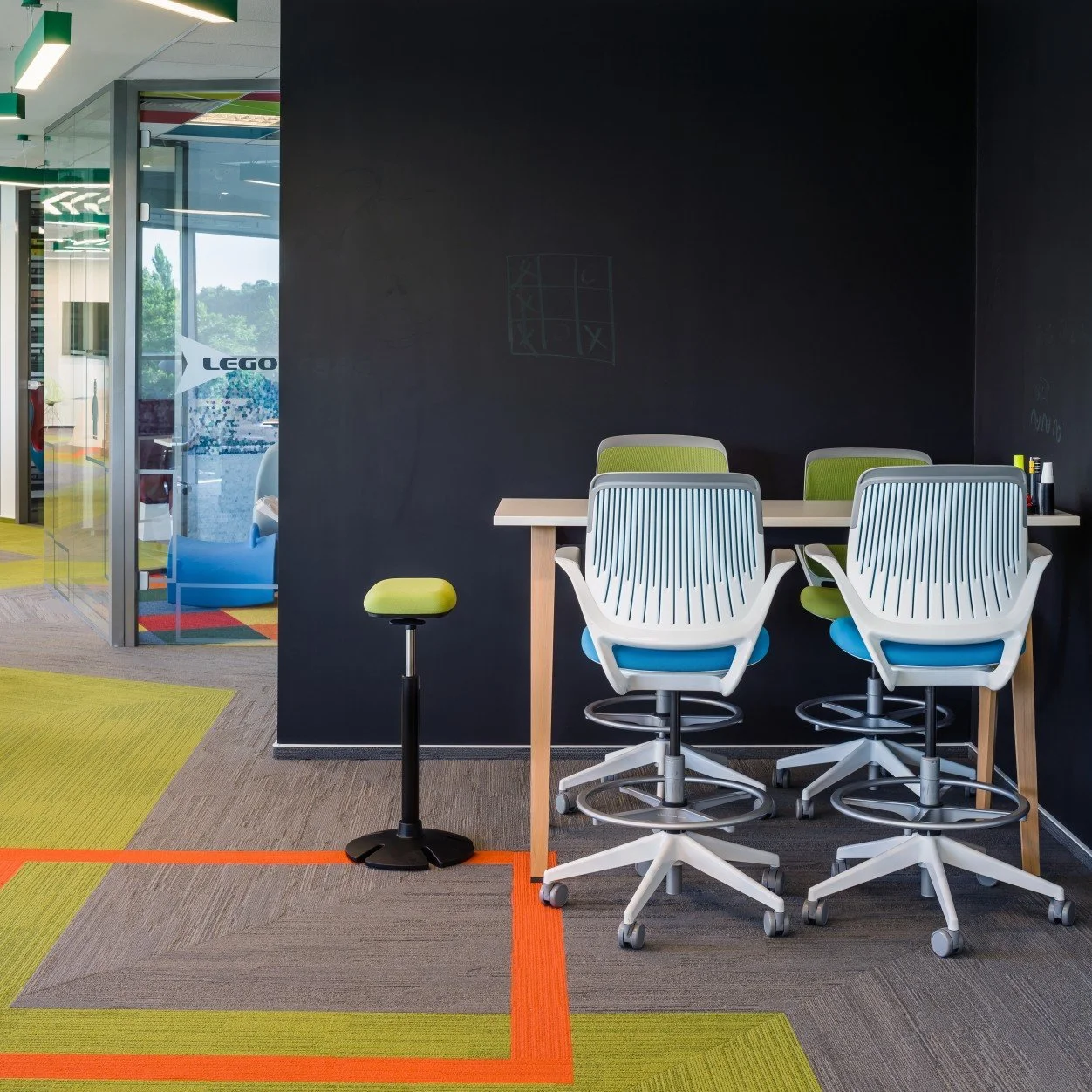 Modern office space with a black accent wall, white ergonomic chairs with green cushions, a light wood table, and colorful multi-colored carpet tiles.