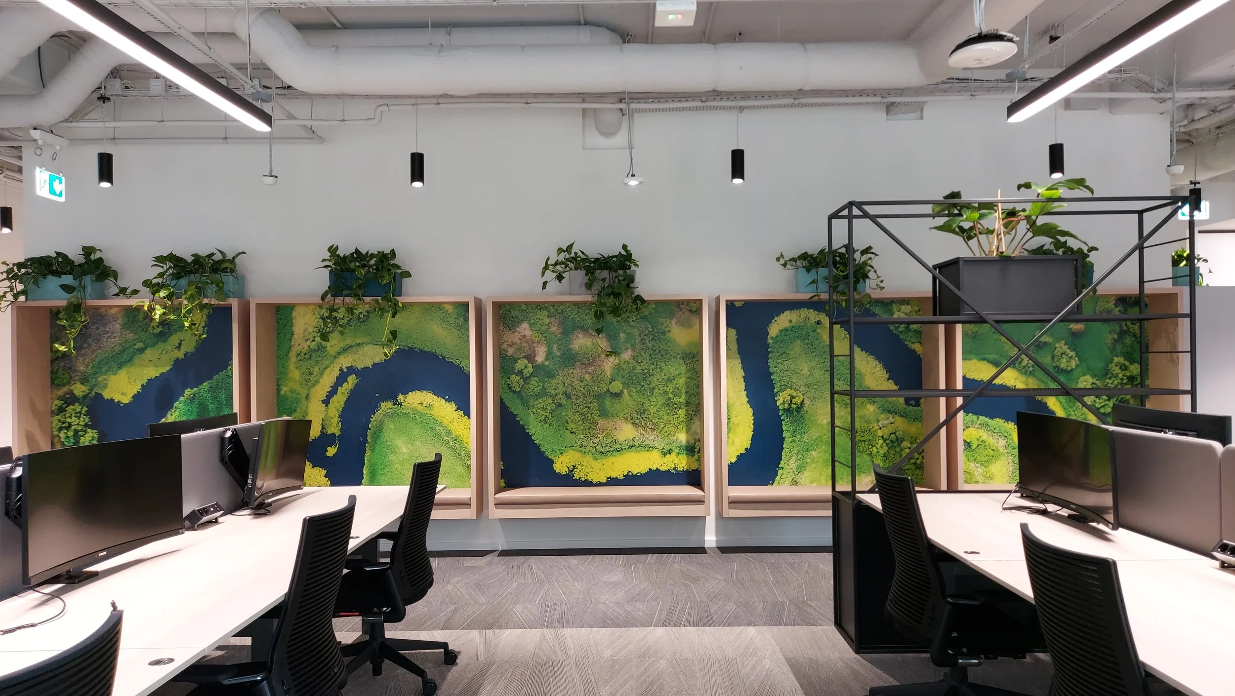 A modern office space with white desks, black chairs, and multiple computer monitors. The background features decorative panels with aerial views of lush green landscapes and winding rivers, topped with potted plants. Overhead lighting and exposed ceiling pipes are visible.