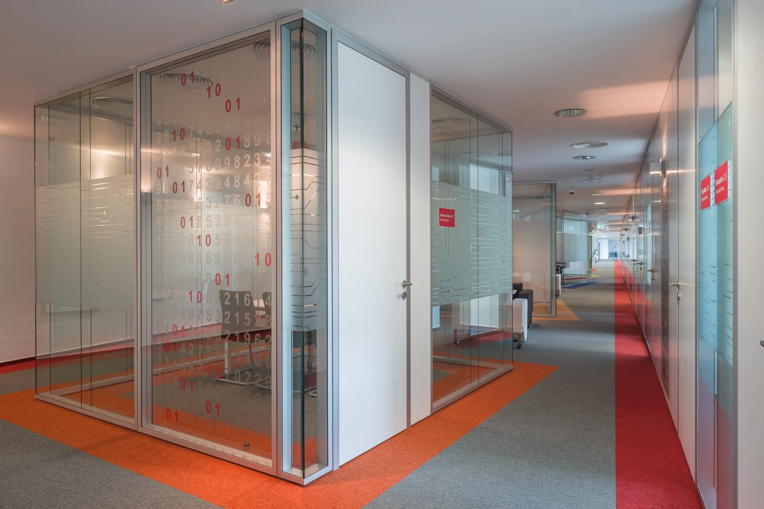 Modern office corridor with glass enclosed rooms, some with red digital data displays, and red signs on the walls, with multicolored carpet flooring.