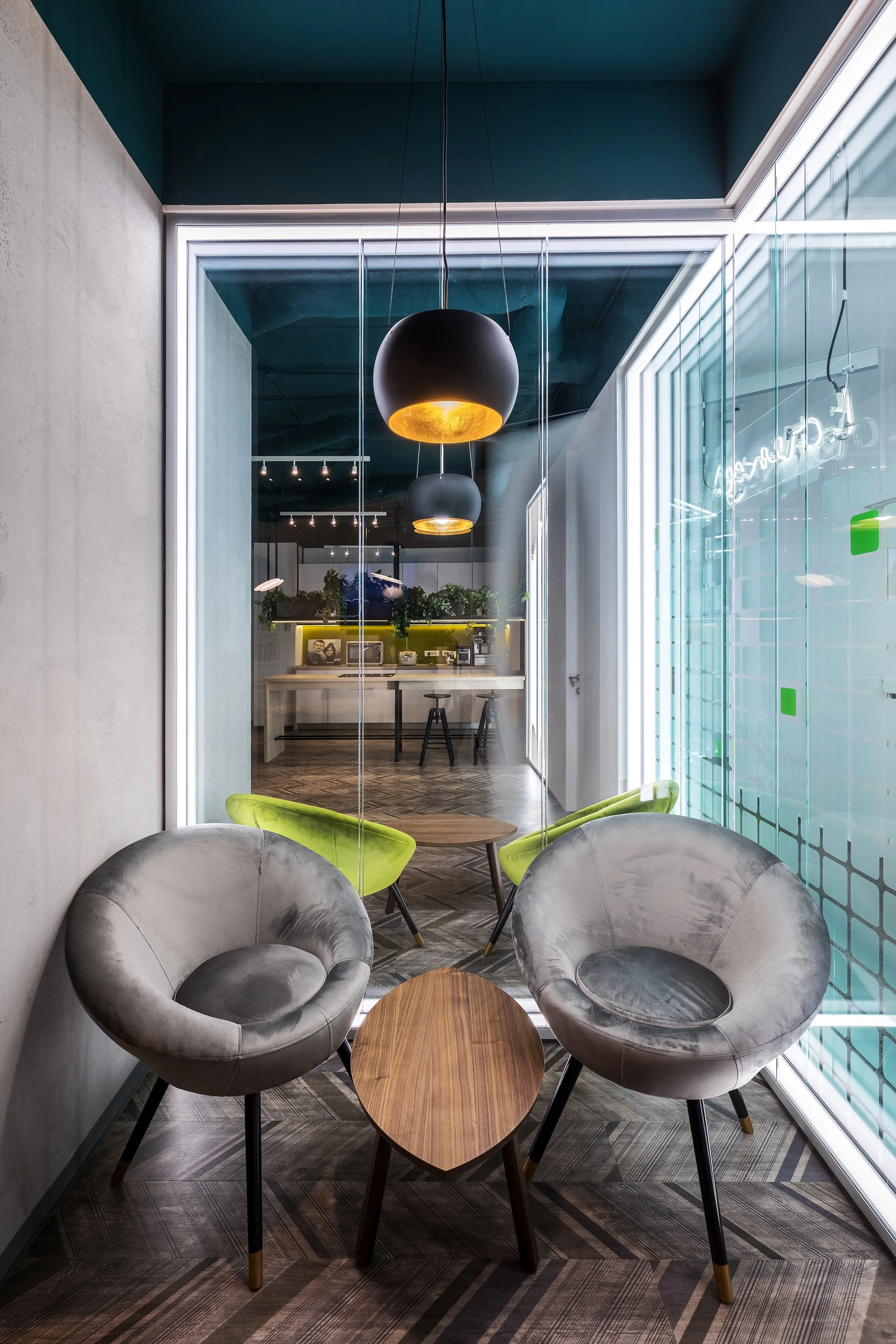 Modern office lounge area with two gray velvet chairs, two green chairs, a small wooden table, and large windows with glass panels. Pendant lights hang from the ceiling.