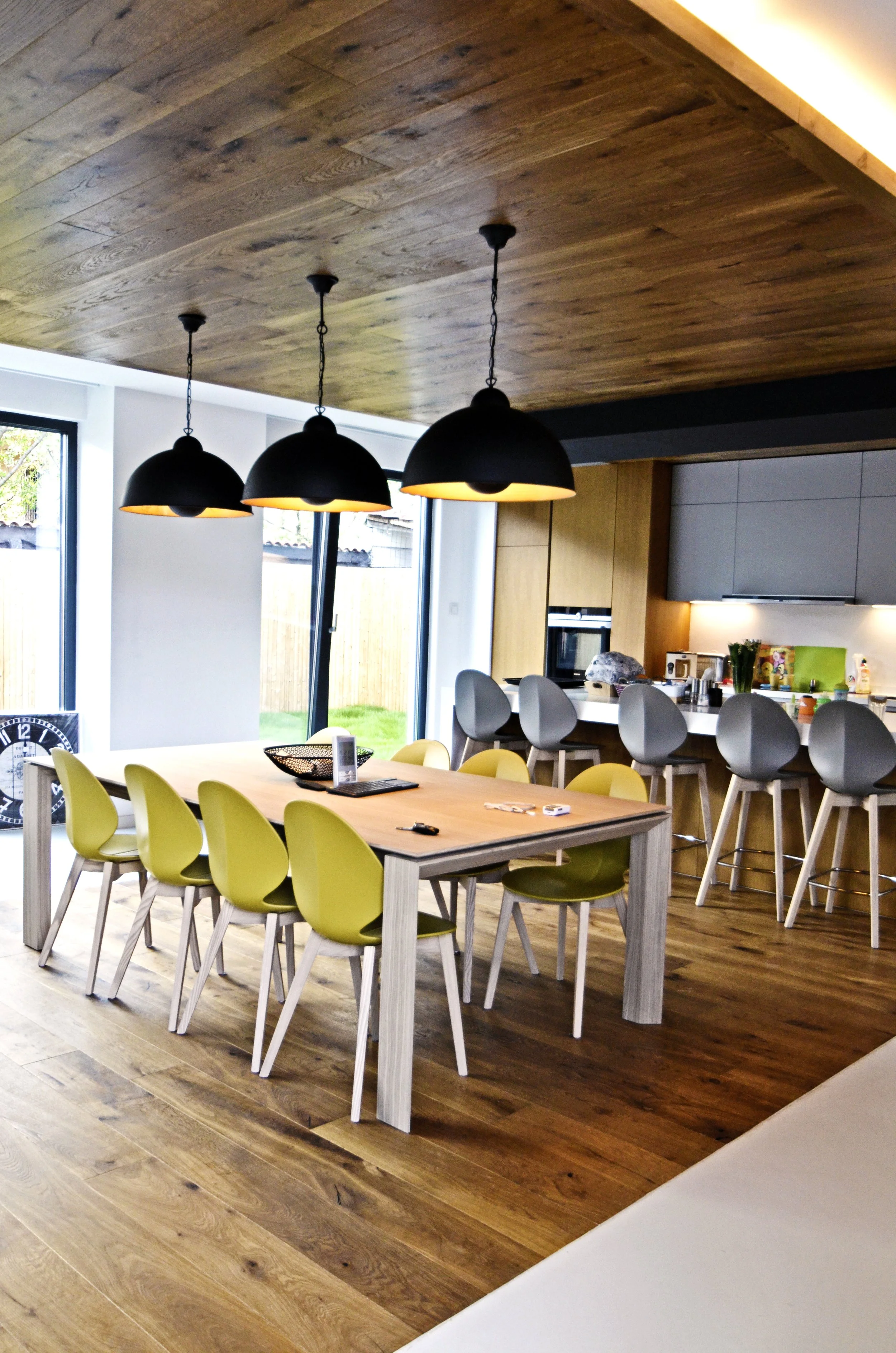 Modern kitchen with a wooden dining table, yellow chairs, black pendant lights, and a bar counter with gray stools.