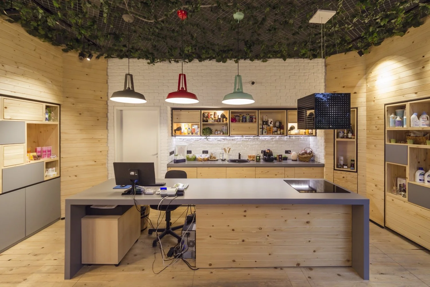 A modern kitchen with wooden paneling, white brick wall, and colorful hanging pendant lights. There is a large kitchen island with a computer and various office supplies, a black stovetop, and open shelving with kitchen items and snacks. The ceiling has decorative greenery.