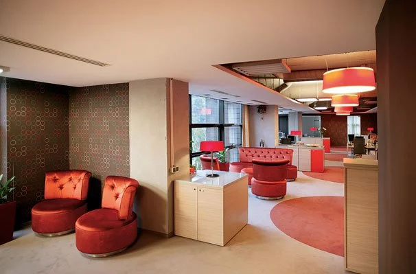 Interior view of a modern lounge or hotel lobby with red upholstered chairs, round ottomans, and contemporary lighting fixtures.