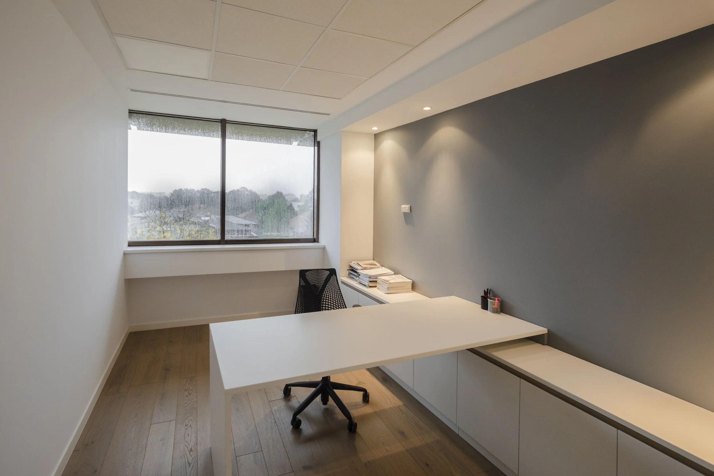 Empty office with a white desk, black mesh chair, window view of trees and buildings, and a gray accent wall with recessed lighting.