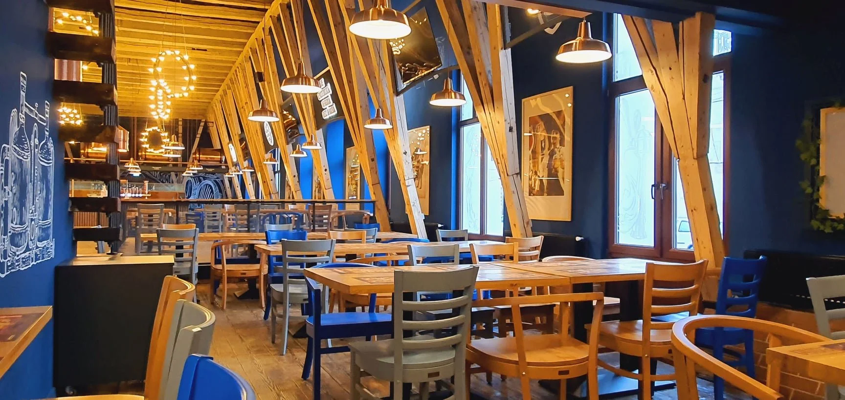 Interior of a cozy restaurant with wooden tables and chairs, Navy blue walls, large windows, and warm pendant lighting, decorated with framed artwork and wall murals.