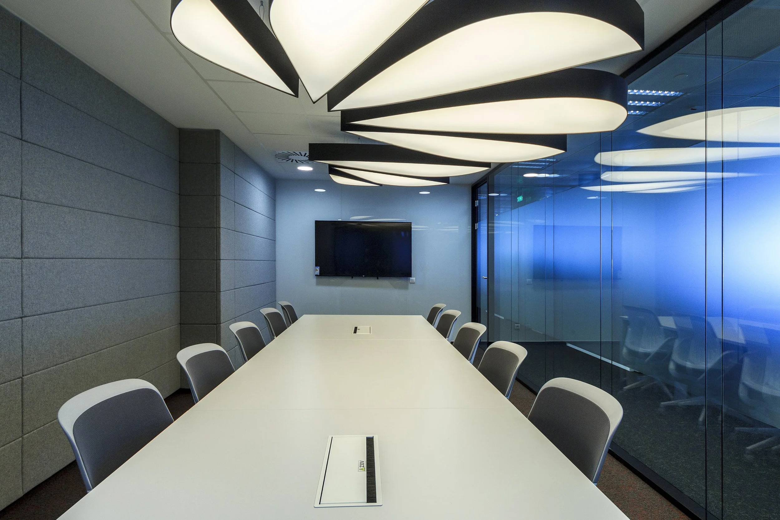 Empty modern conference room with white table, chairs, large ceiling lights, a wall-mounted TV, gray padded wall, and glass walls with blue tint.
