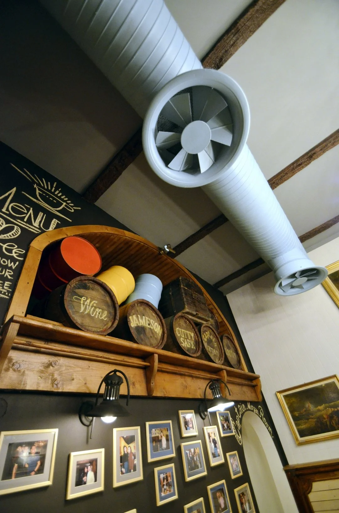 Ceiling-mounted white industrial exhaust fan in a restaurant or pub, with wooden barrels labeled 'Wine,' 'Jameson,' 'City Sark,' and others displayed on a high shelf below, framed photographs on a black wall, and a painting in a gold frame.