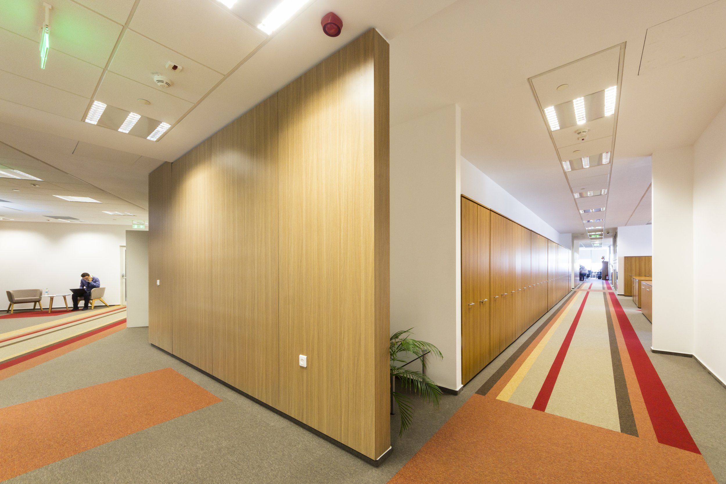 A hallway in a modern office building with wood-paneled walls, a colorful striped carpet, and ceiling lights. A man is sitting on a bench in the waiting area to the left.