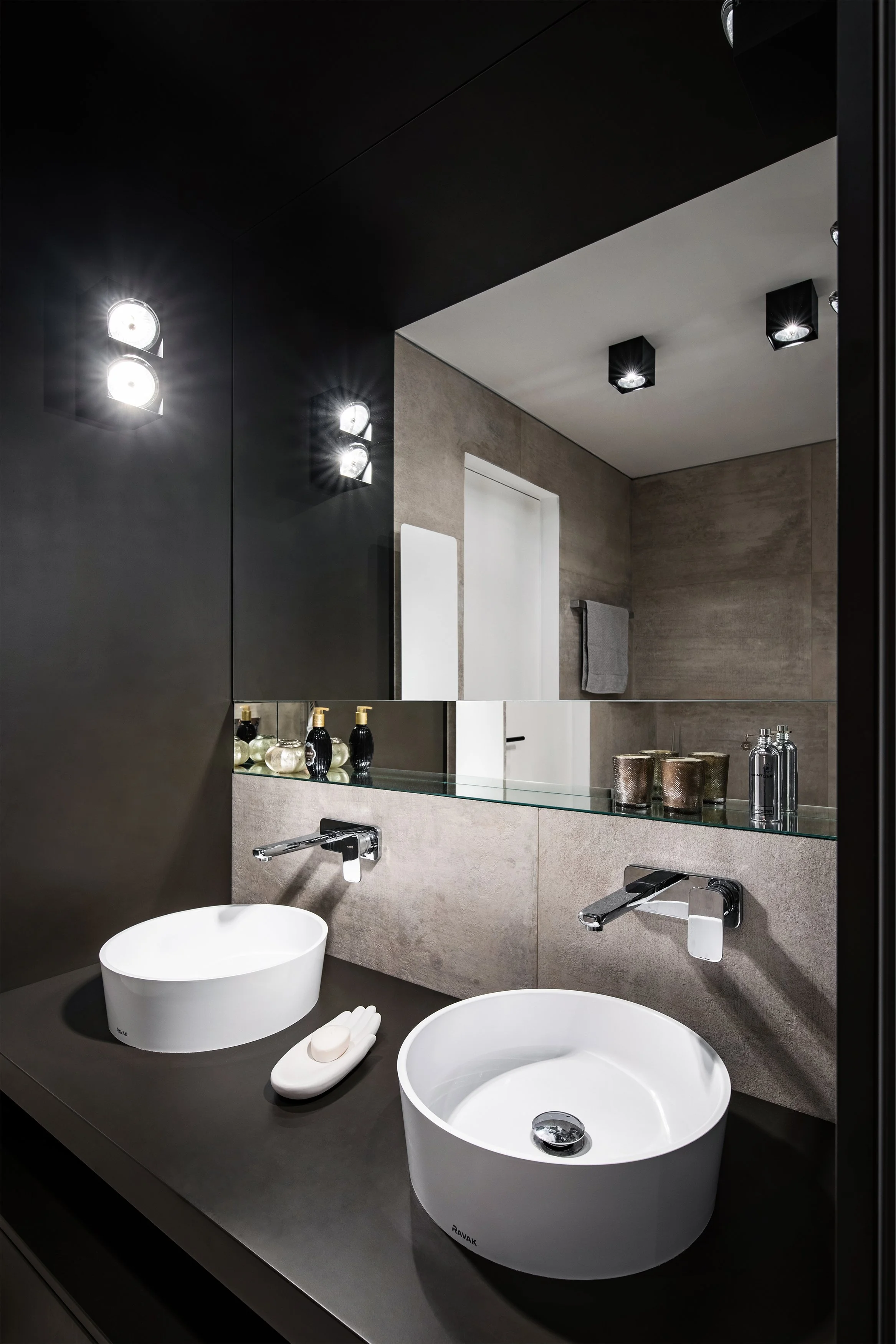 Modern bathroom with double vessel sinks, large mirror, wall-mounted faucets, and stylish decor items. Dark accents and contemporary lighting fixtures.