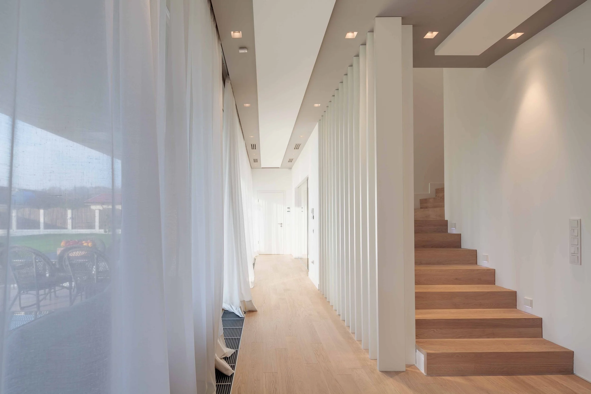 Modern interior hallway with wooden stairs on the right, white walls, ceiling lights, and sheer white curtains on the left with sunlight coming through.