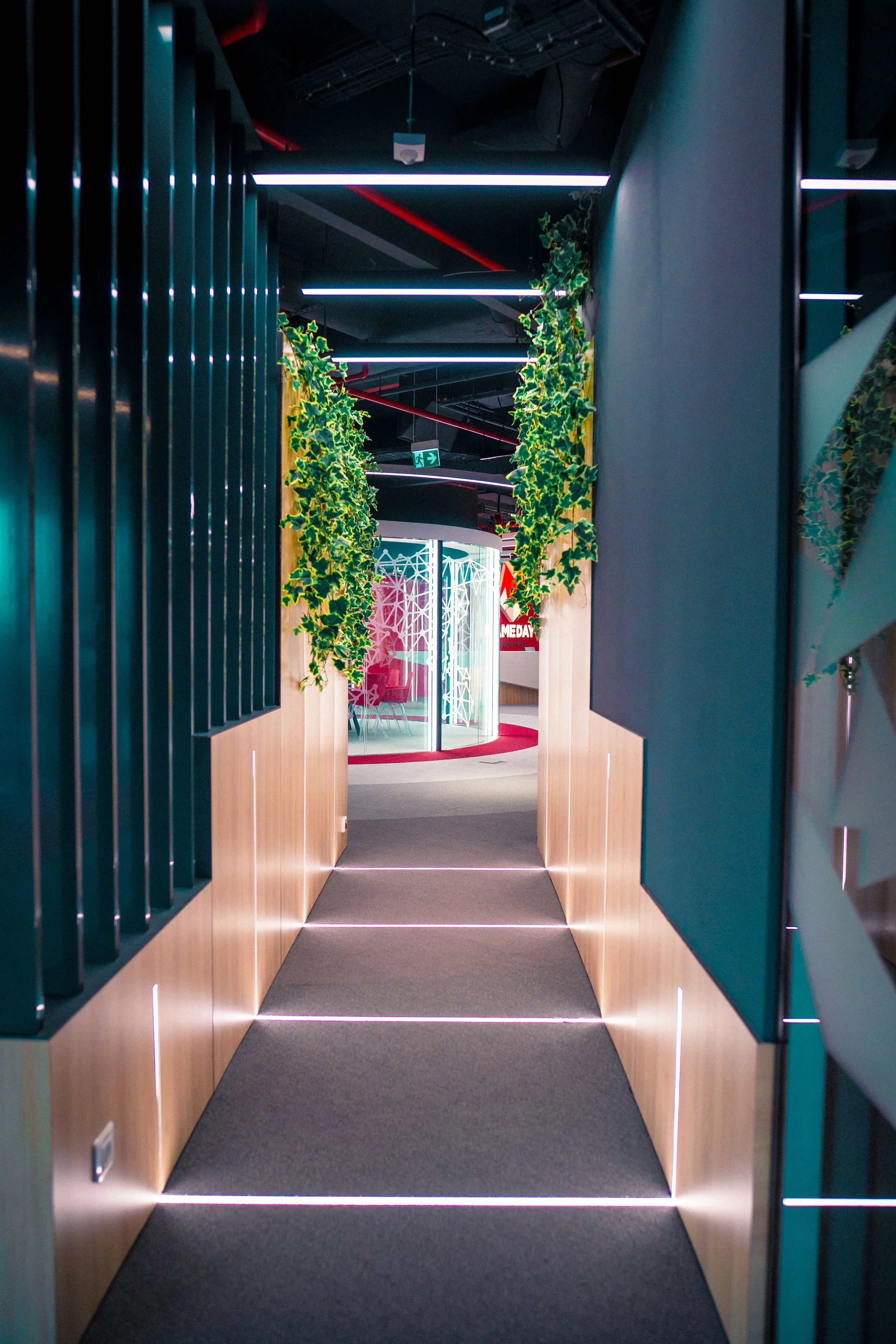 Modern corridor with plants on both sides, leading to a circular reception or meeting area with vibrant pink seating and colorful geometric decor.