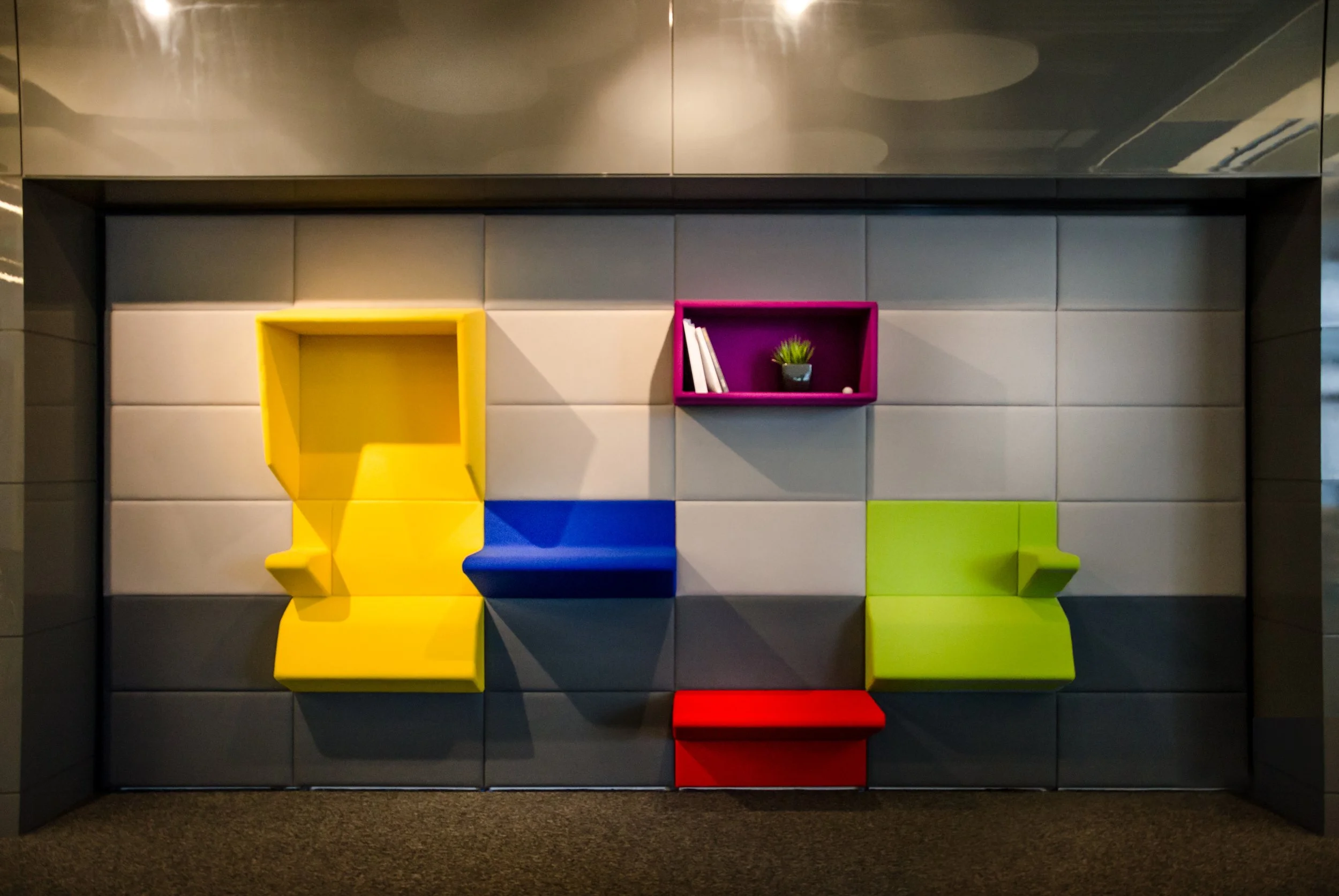 Colorful modern seating wall with yellow, blue, green, and red molded seats, and pink and purple cubed shelves with books and a small plant, against a tiled wall and carpeted floor.