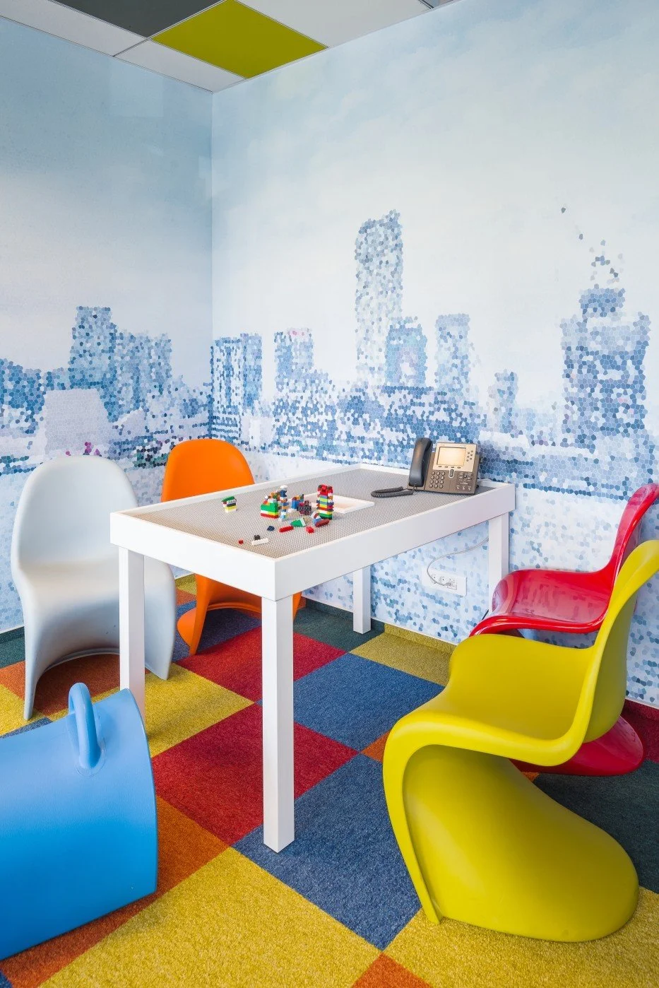 Colorful modern office room with mosaic cityscape mural, a white table with building blocks, and bright, uniquely shaped chairs in yellow, red, blue, orange, and white on multicolored checkered carpet.