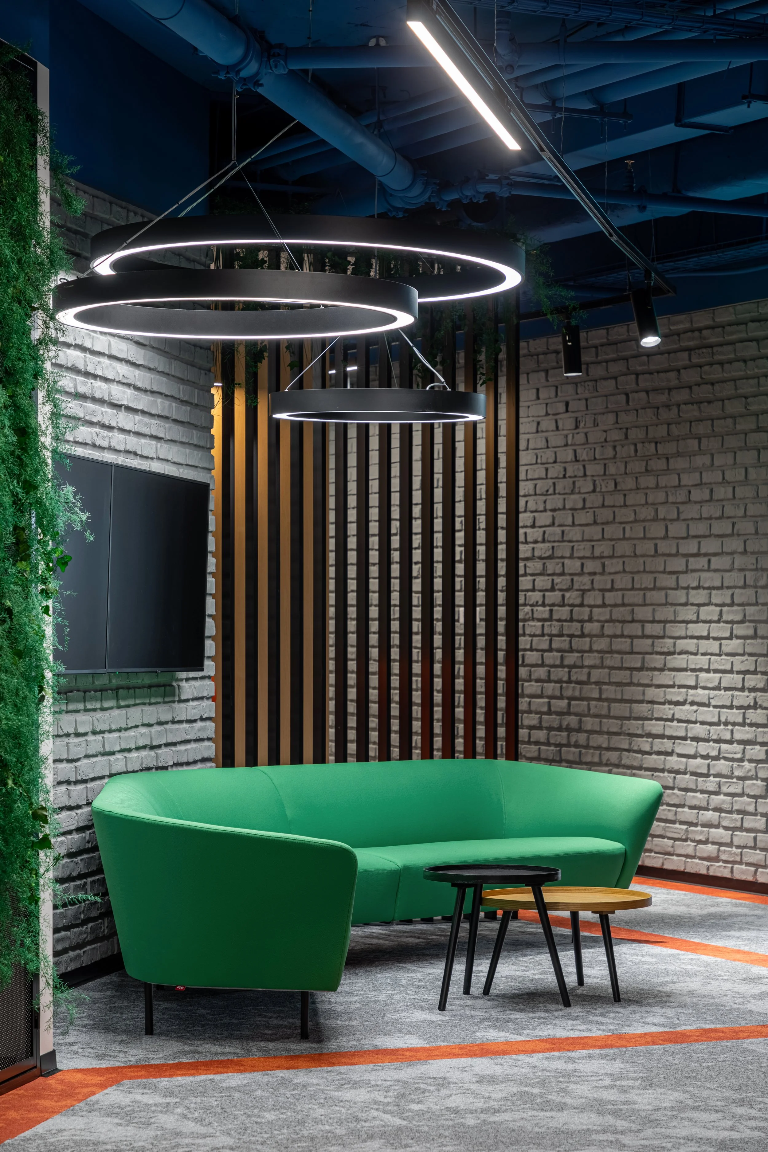 Modern lounge area with a bright green sofa, two small nesting tables, decorative green plants, wall-mounted TV, and contemporary lighting fixtures.