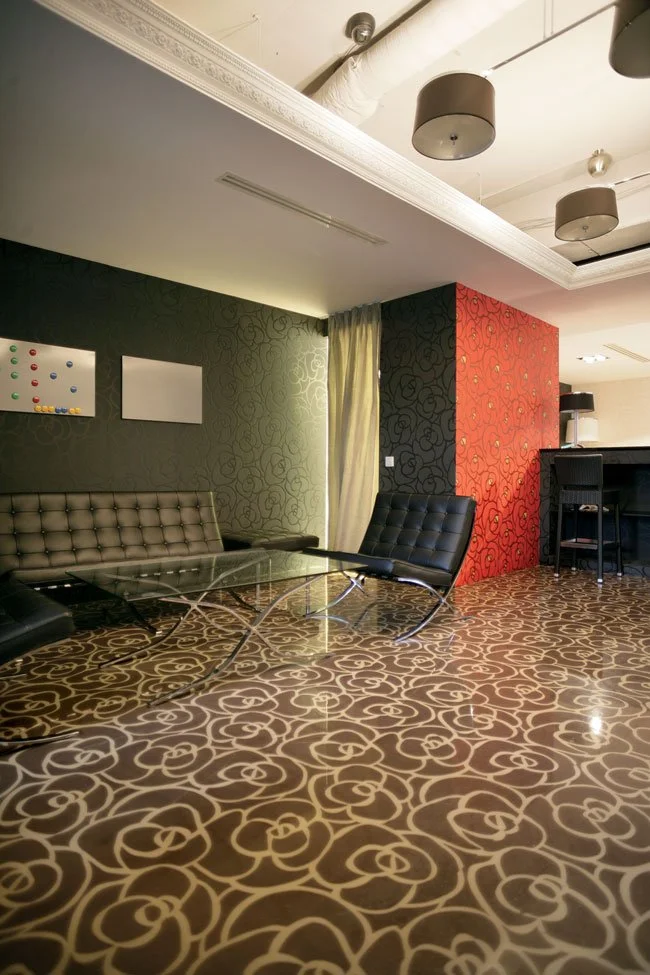Modern lounge area with patterned carpet, black and beige chairs, glass coffee table, decorative patterned walls, curtain, and ceiling lights.