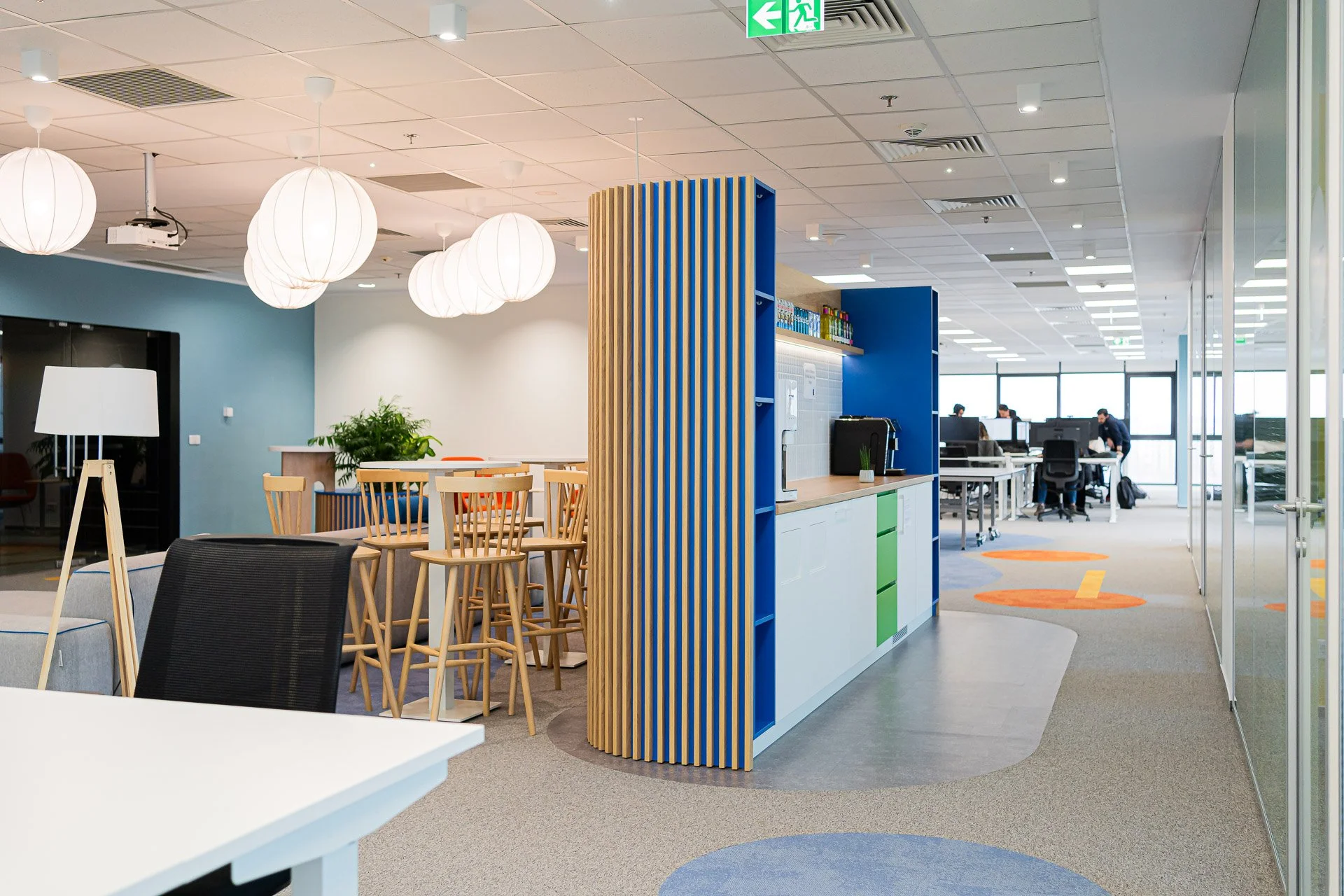 Modern office break room with seating, a kitchenette, and workstations in the background.