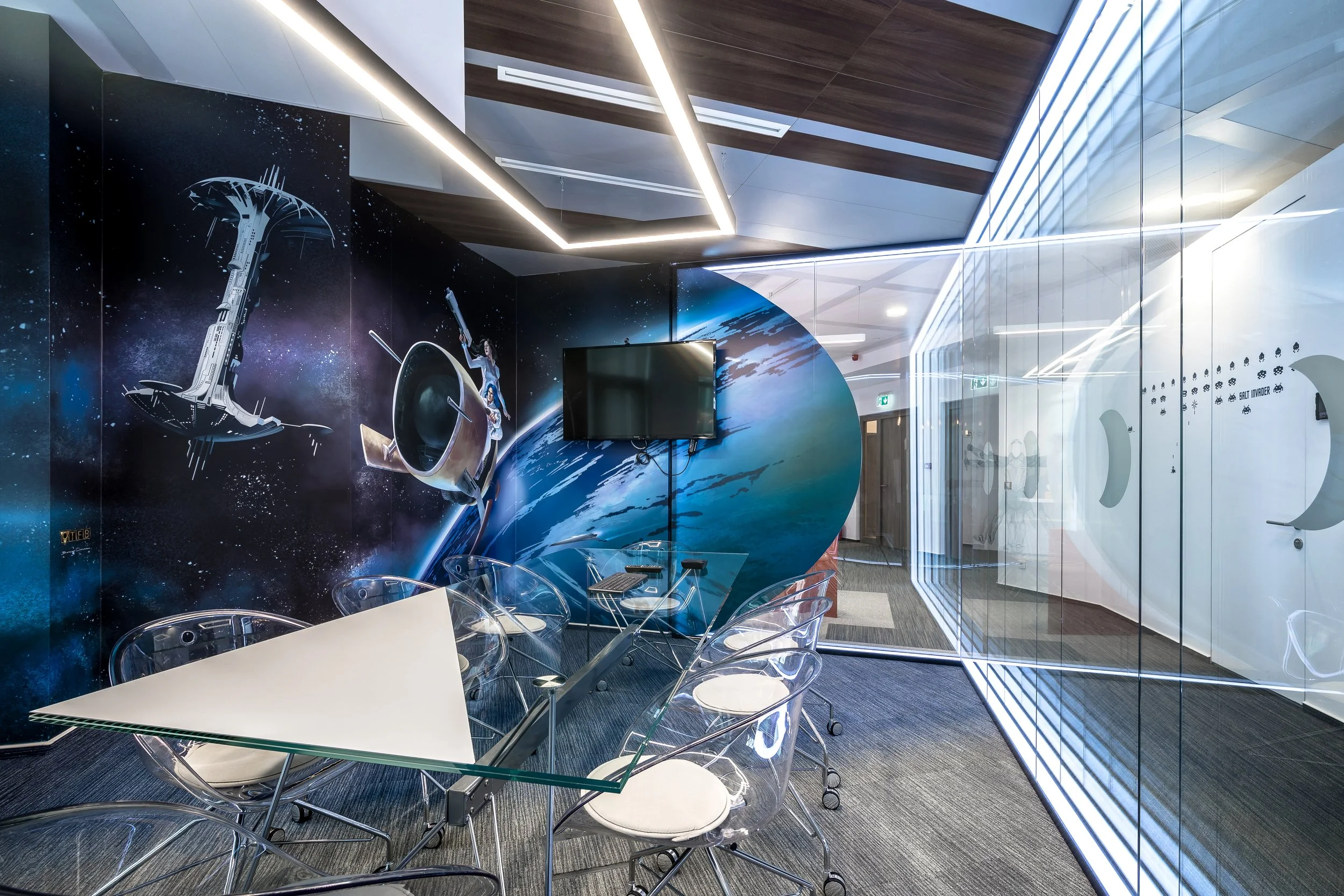 A modern office conference room with space-themed wall art, a glass table with transparent chairs, a wall-mounted TV, and a glass wall with futuristic spaceship designs and moon phases.