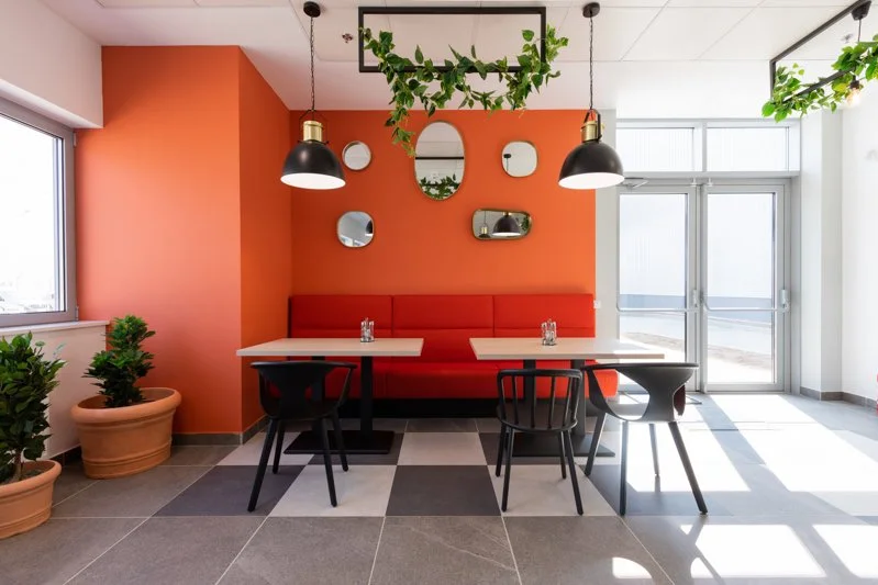Modern dining area with orange accent wall, red cushioned bench, two white tables, black chairs, hanging black pendant lights, potted plants, and wall mirrors, with large windows and glass door allowing natural light.