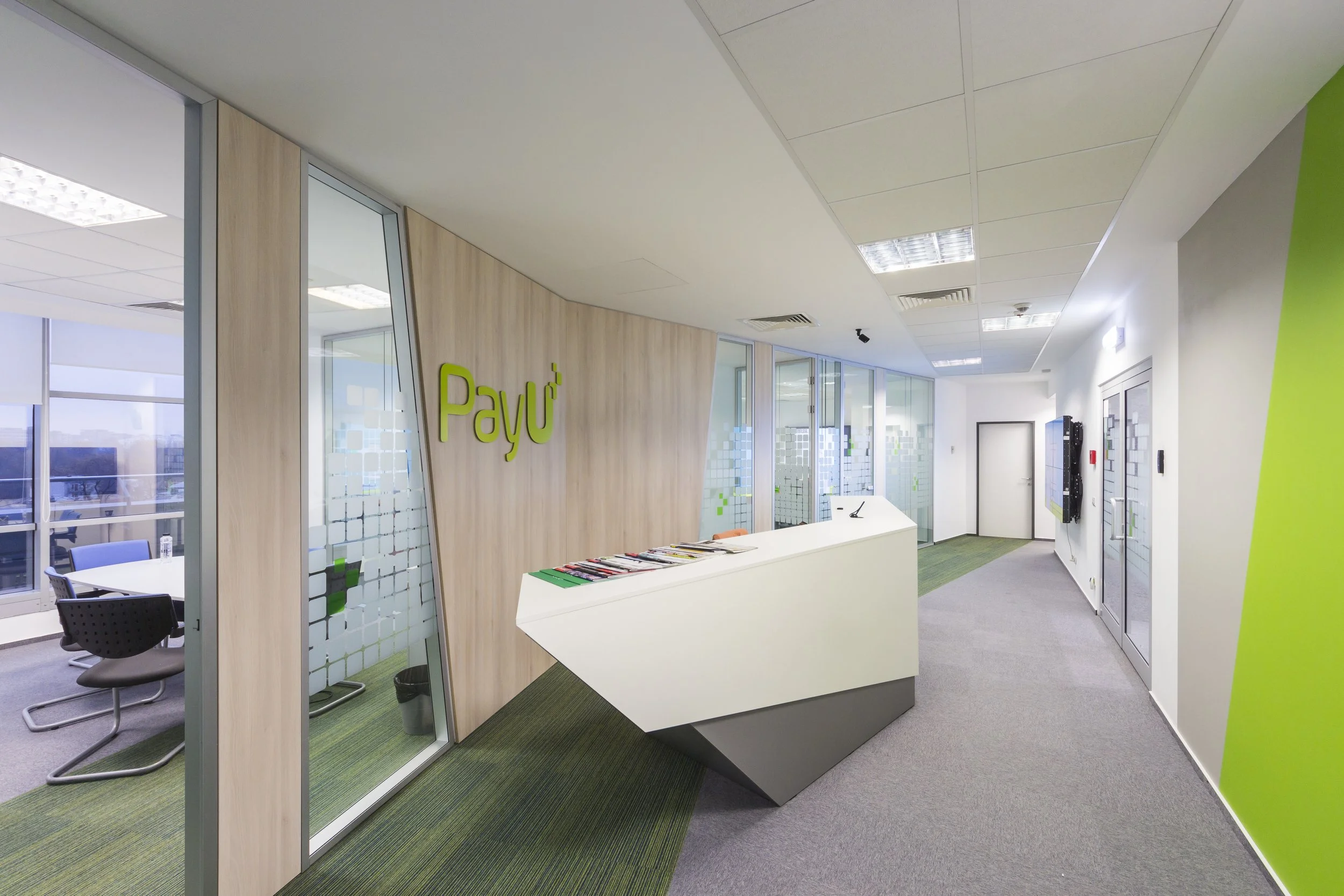 Modern office reception area with a white angular desk, glass conference room doors with frosted squares, green and gray color scheme, and the company logo 'Payu' on the wall.
