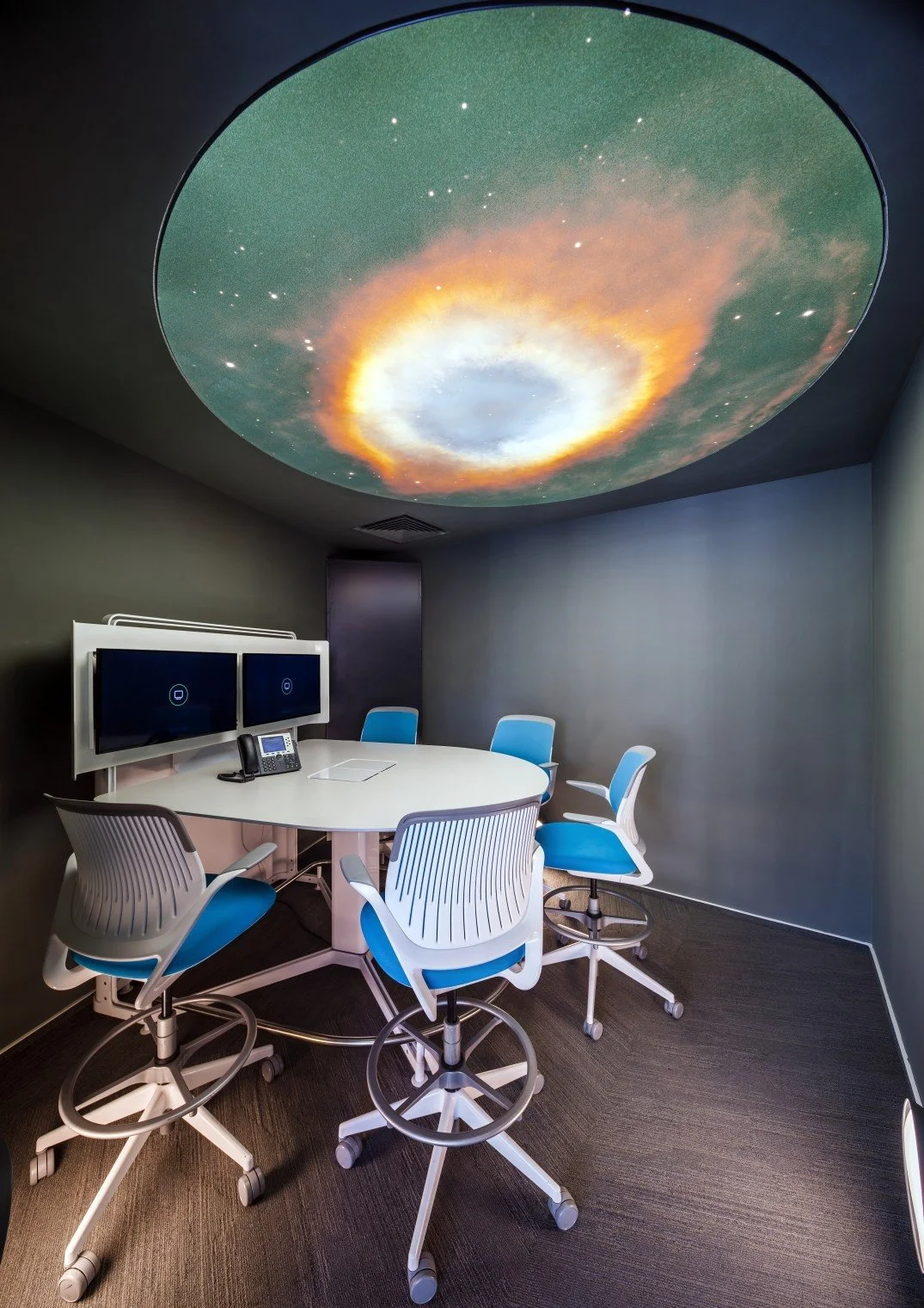 A small conference room with a round white table, five white and blue office chairs, two monitors, a telephone, and a ceiling projection of a galaxy or nebula on the ceiling.