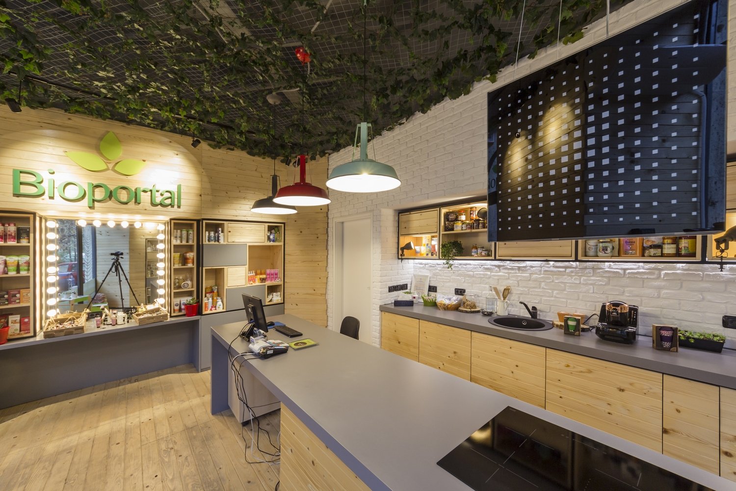 Interior of a modern office space with a kitchen area, wooden cabinets, a desk with a computer, and a large television screen. The space features a white brick wall, ceiling greenery, hanging colorful lamps, and the sign 'Biportal' with a leaf logo.