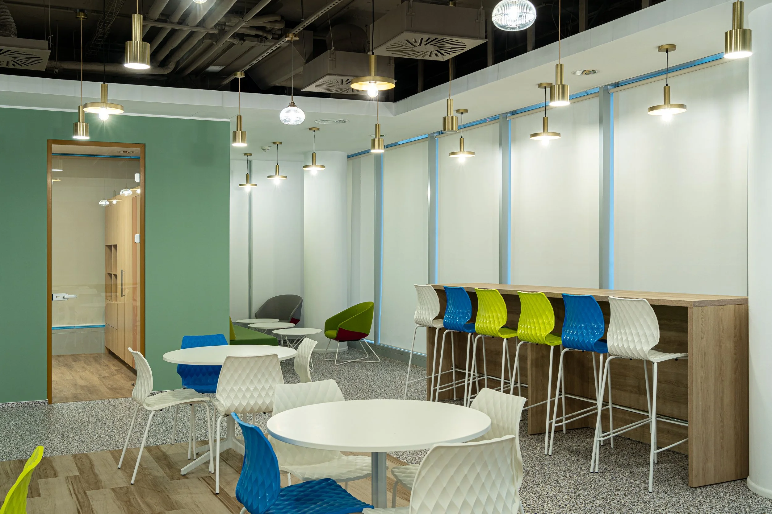 Modern coworking space with colorful chairs, white tables, a bar-height counter with stools, and decorative pendant lighting.
