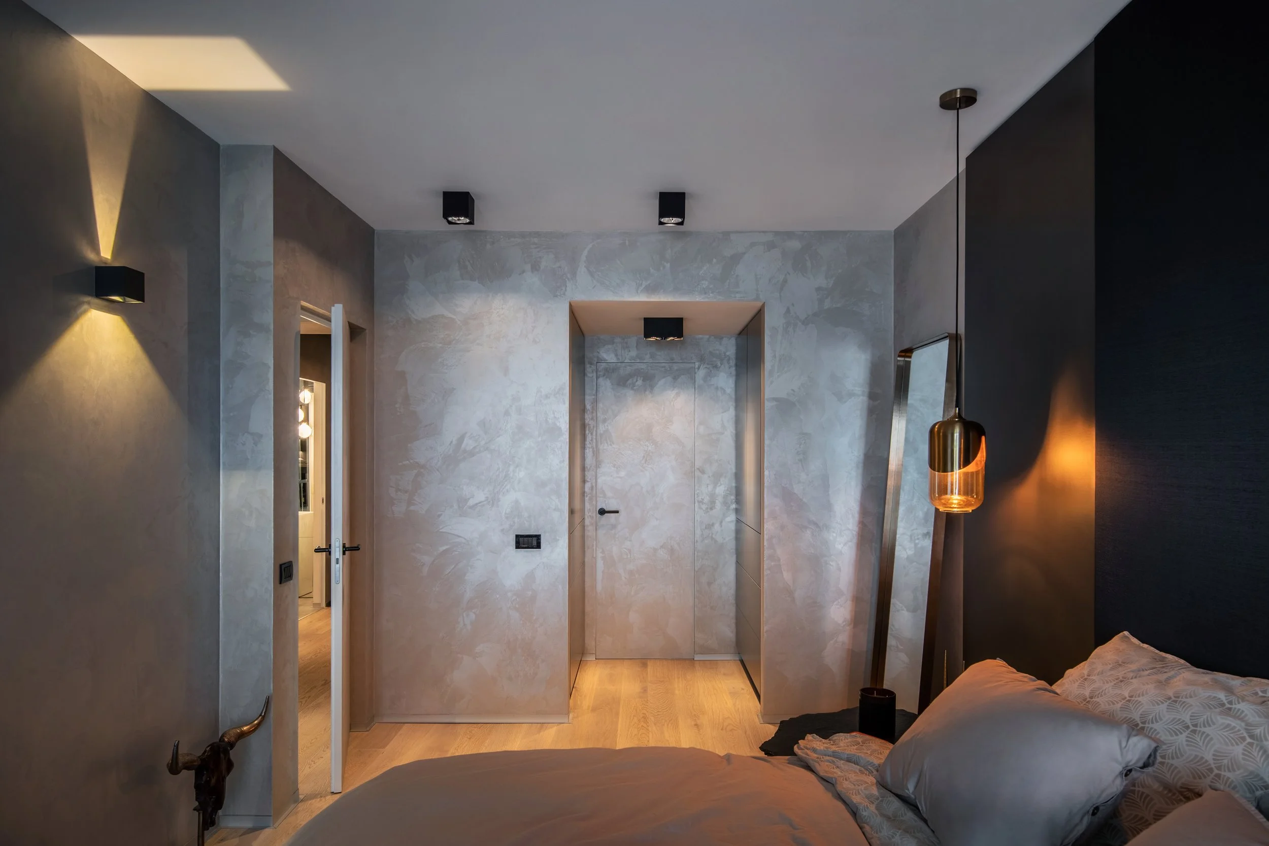 Modern bedroom with textured gray walls, a bed with beige bedding, a hanging pendant light, a large mirror, and a door at the end of the room.