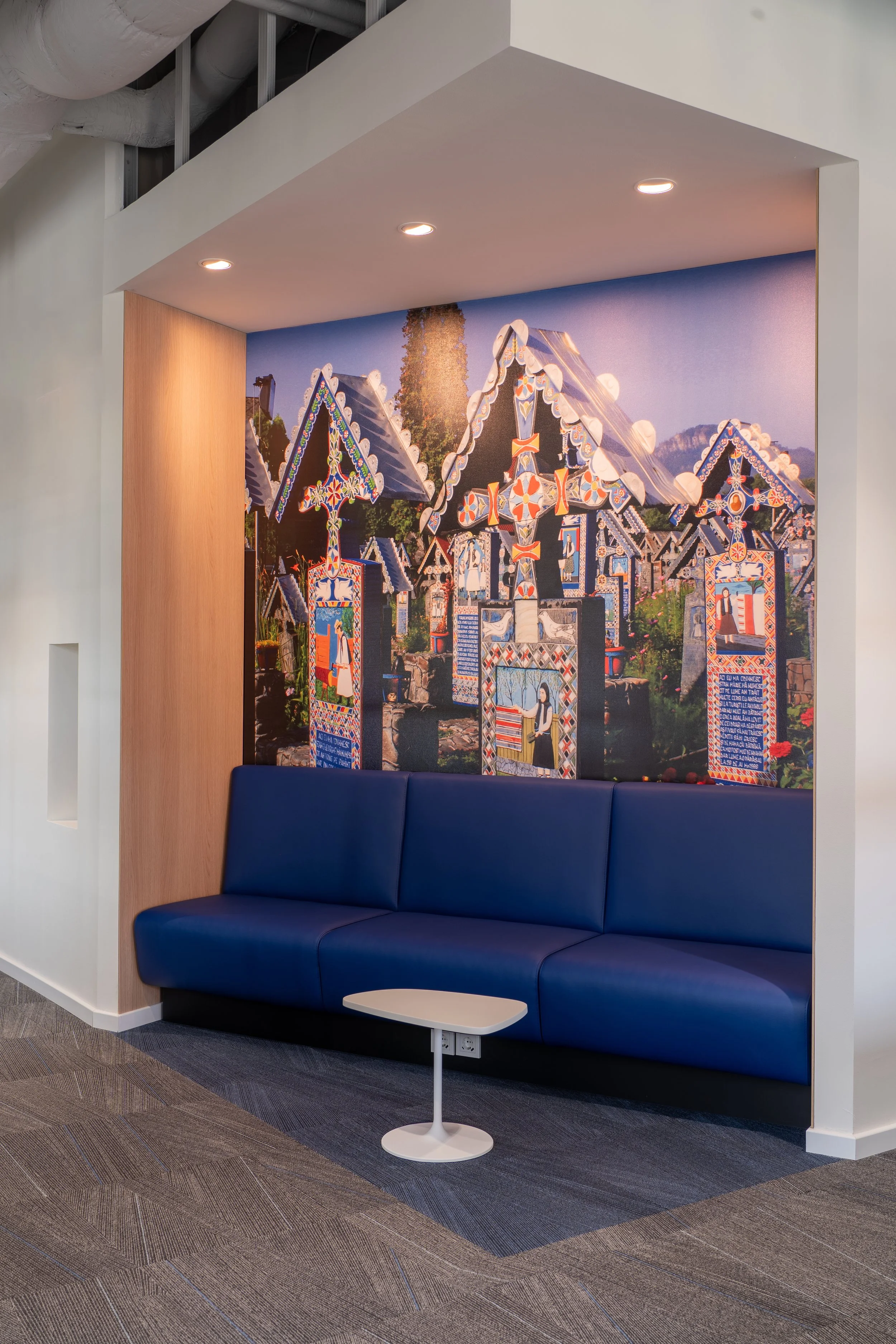 A wall mural of a traditional village scene with decorated buildings and figures in folk costumes, mounted above a blue sofa with a small white table in front of it.