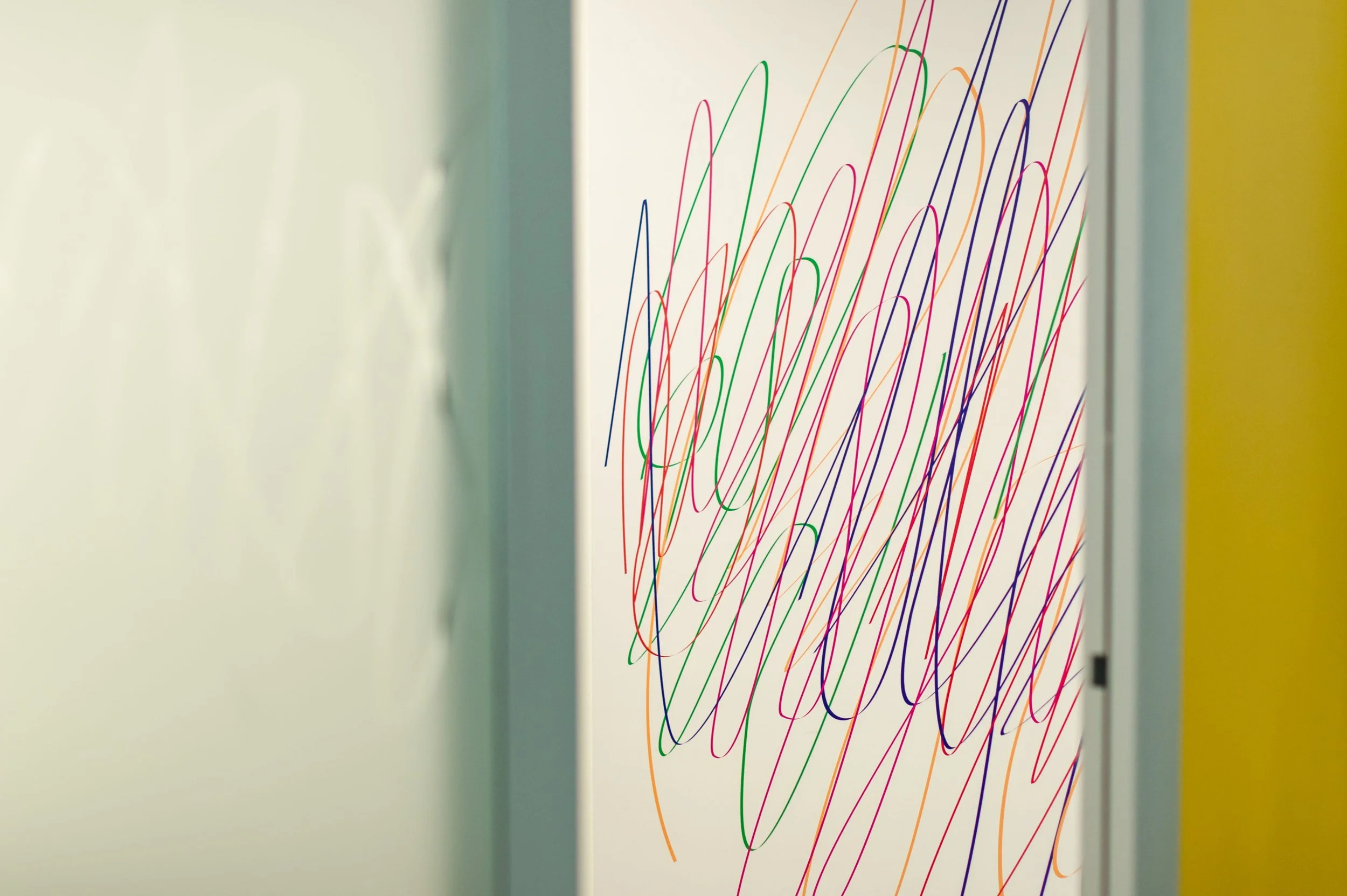 Colorful scribbles on a white wall, viewed through an open door in a hallway with light-colored walls.