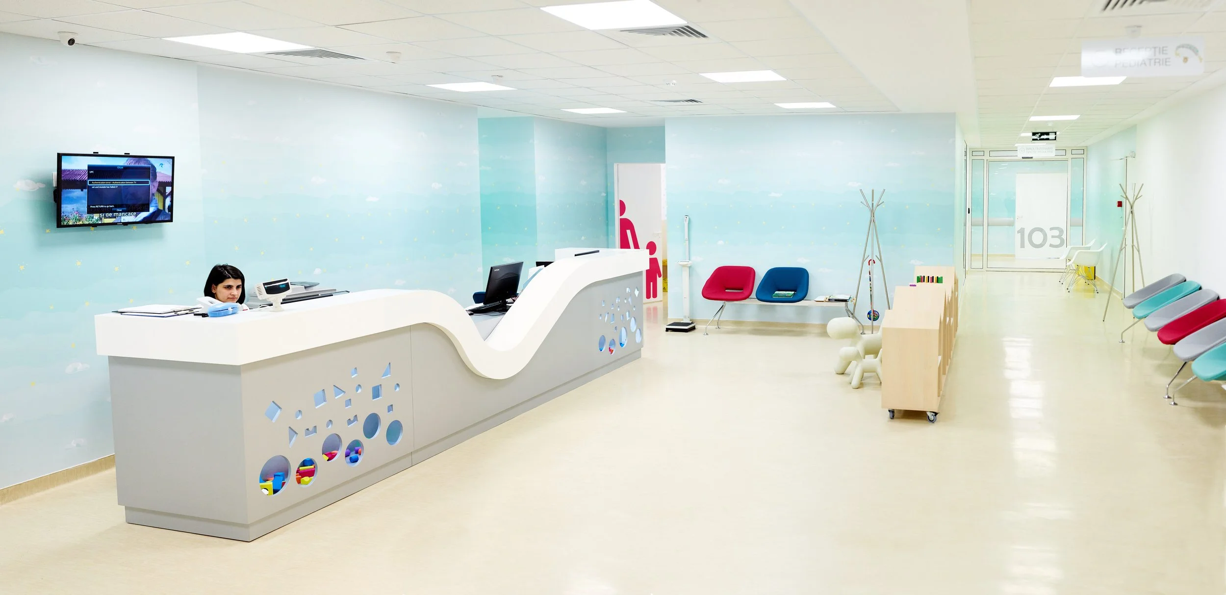 Empty pediatric waiting room with a reception desk, colorful chairs, and play area, decorated with a sky theme with clouds and stars.