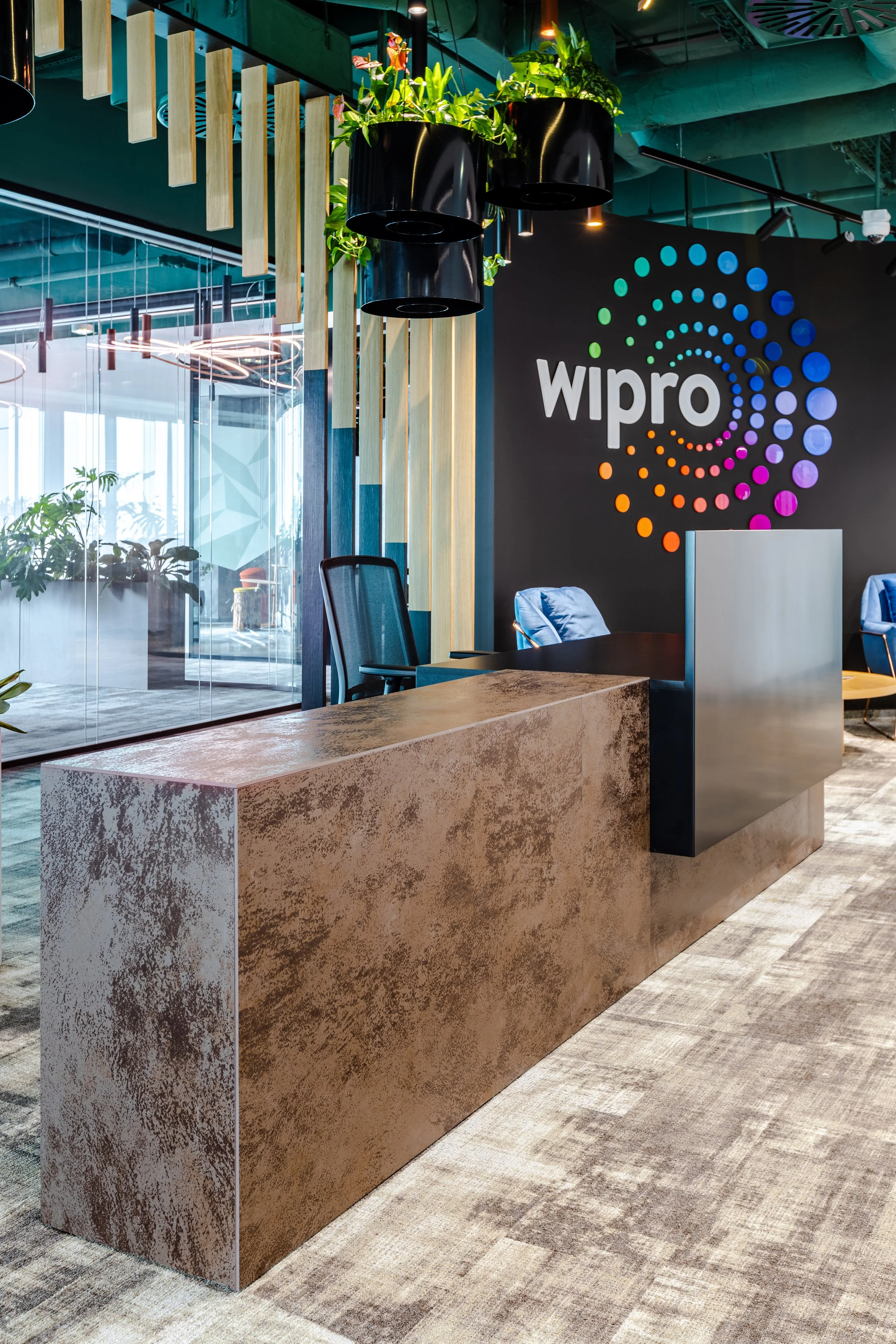 Office reception area with a modern design, featuring a brown textured reception desk and a black wall with a colorful 'wipro' logo, potted plants hanging from the ceiling, and a glass partition in the background.