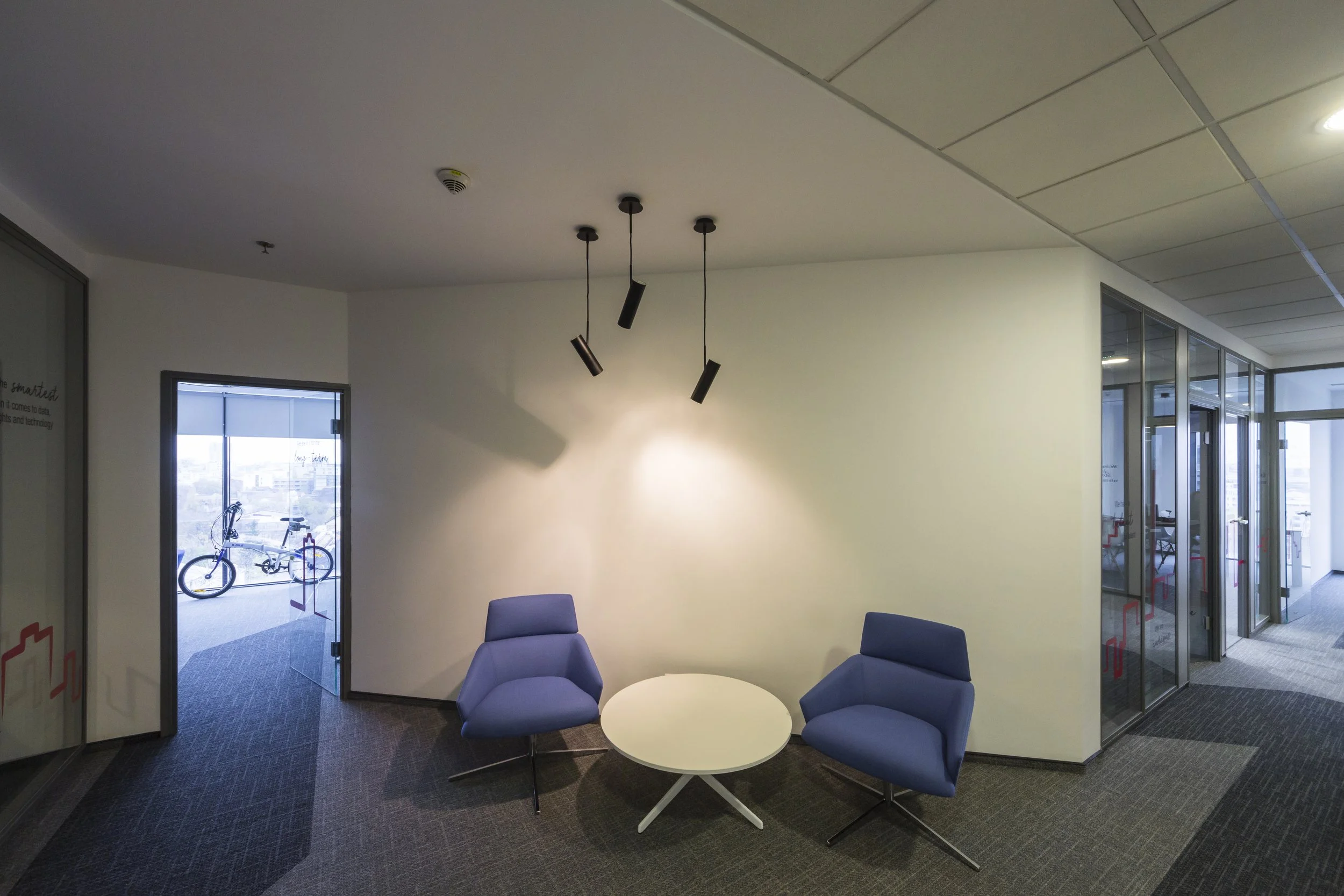 Interior of an office lobby or waiting area with two blue armchairs, a round white table, and modern black ceiling light fixtures. Glass walls and a door leading outside, where a bicycle is visible.