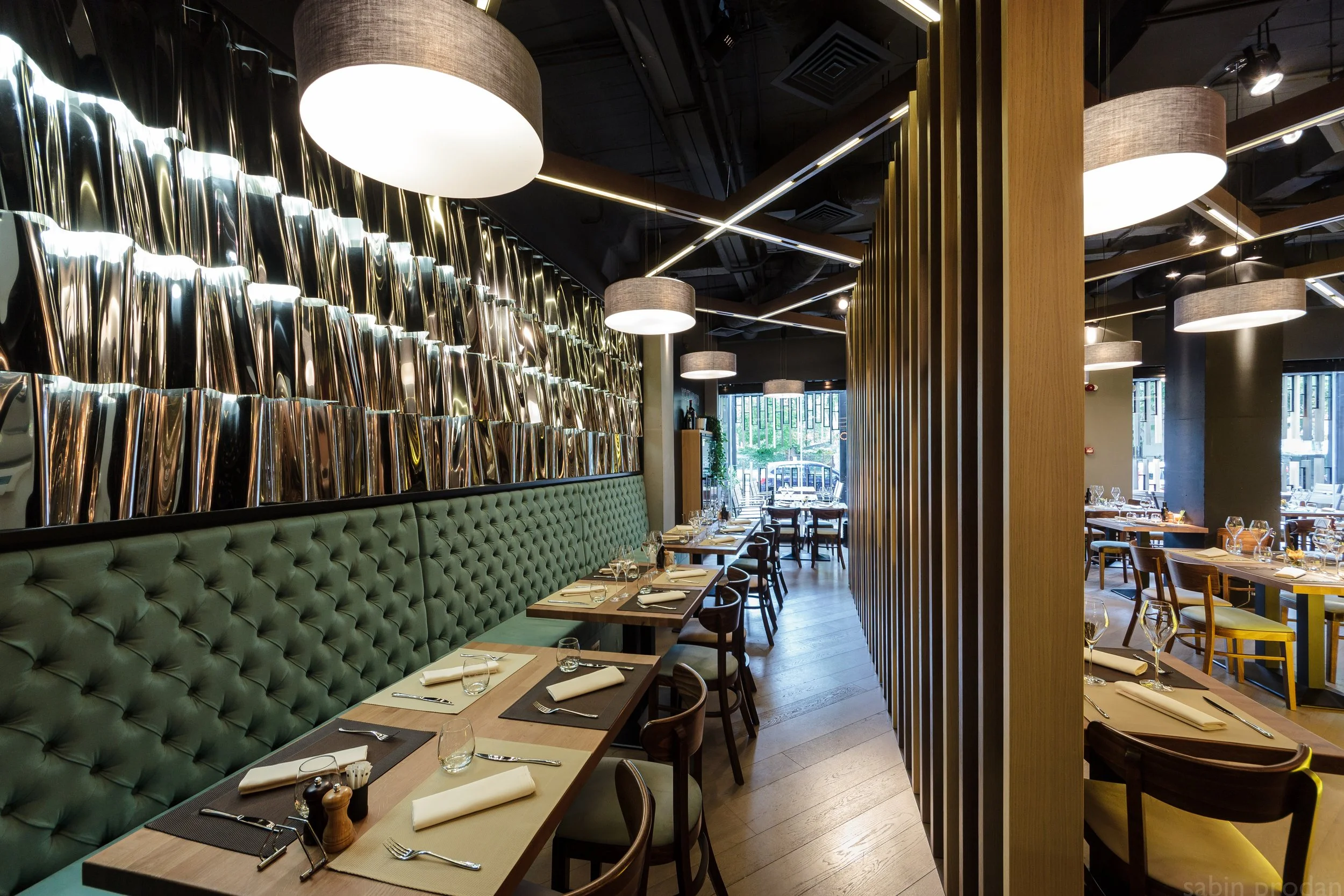 Interior of a modern restaurant with green tufted banquette seating along a wall, wooden divided wall, tables set with napkins, cutlery, glasses, and salt and pepper shakers, large circular hanging lights, and large windows letting in natural light.
