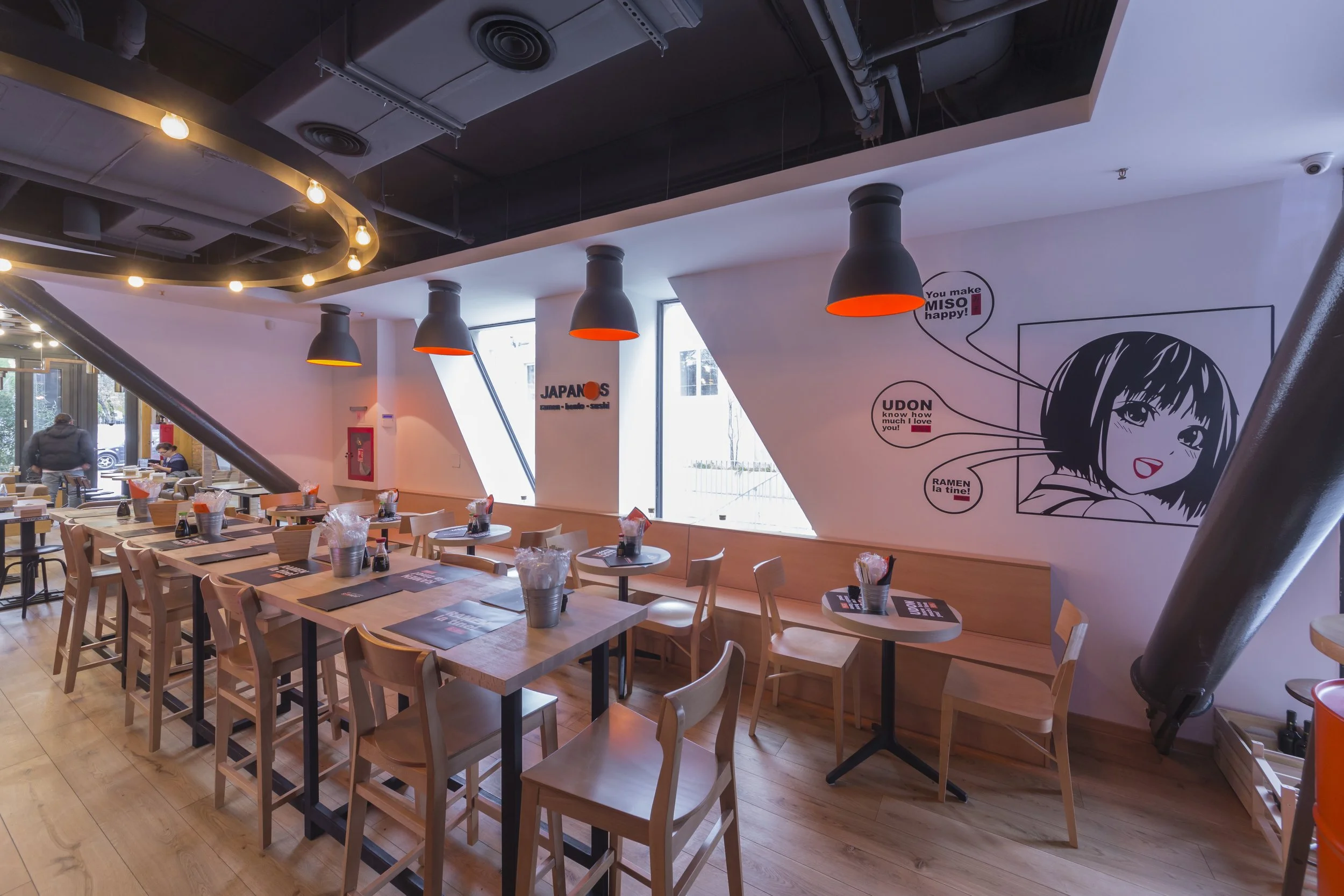 Empty Japanese-themed restaurant with wooden chairs and tables, decorative lighting, and wall art featuring Japanese anime-style illustration with speech bubbles.