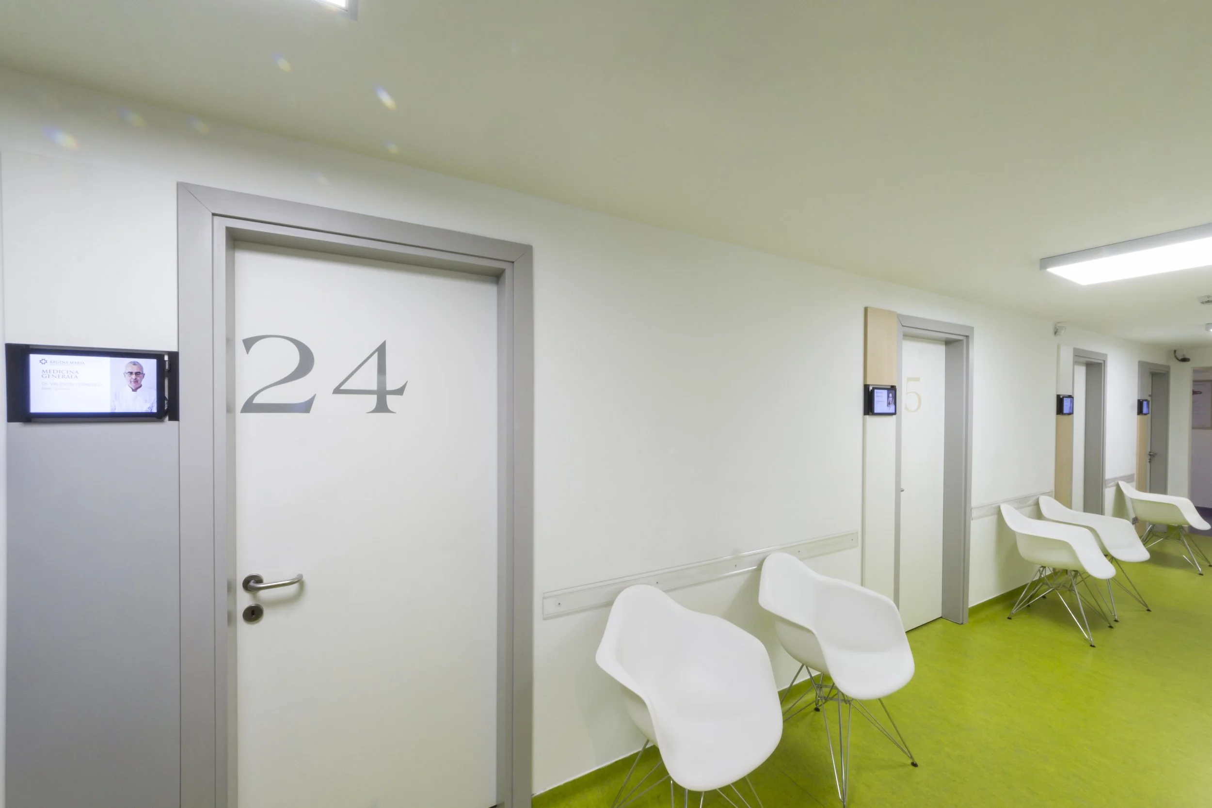 Hospital corridor with doors numbered 24 and 25, white chairs, and wall-mounted screens.