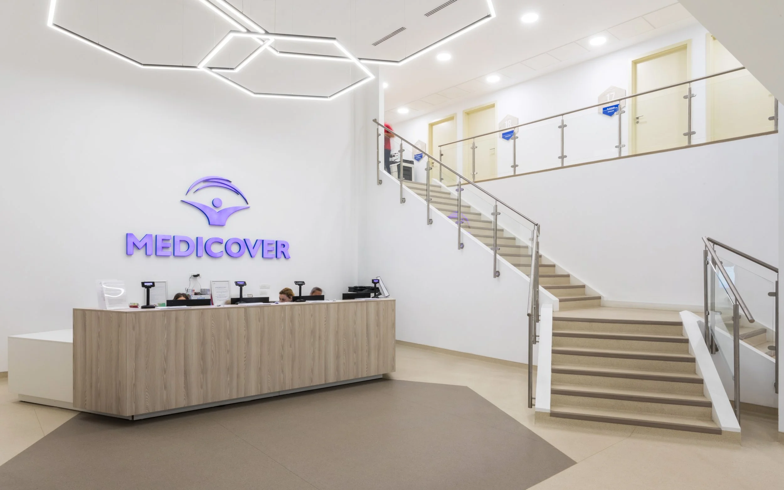 Modern medical clinic reception area with a wooden desk, staff members, and a large illuminated logo on the white wall reading 'MEDICOVER'. A staircase with glass and metal railing leads to an upper floor with closed doors.