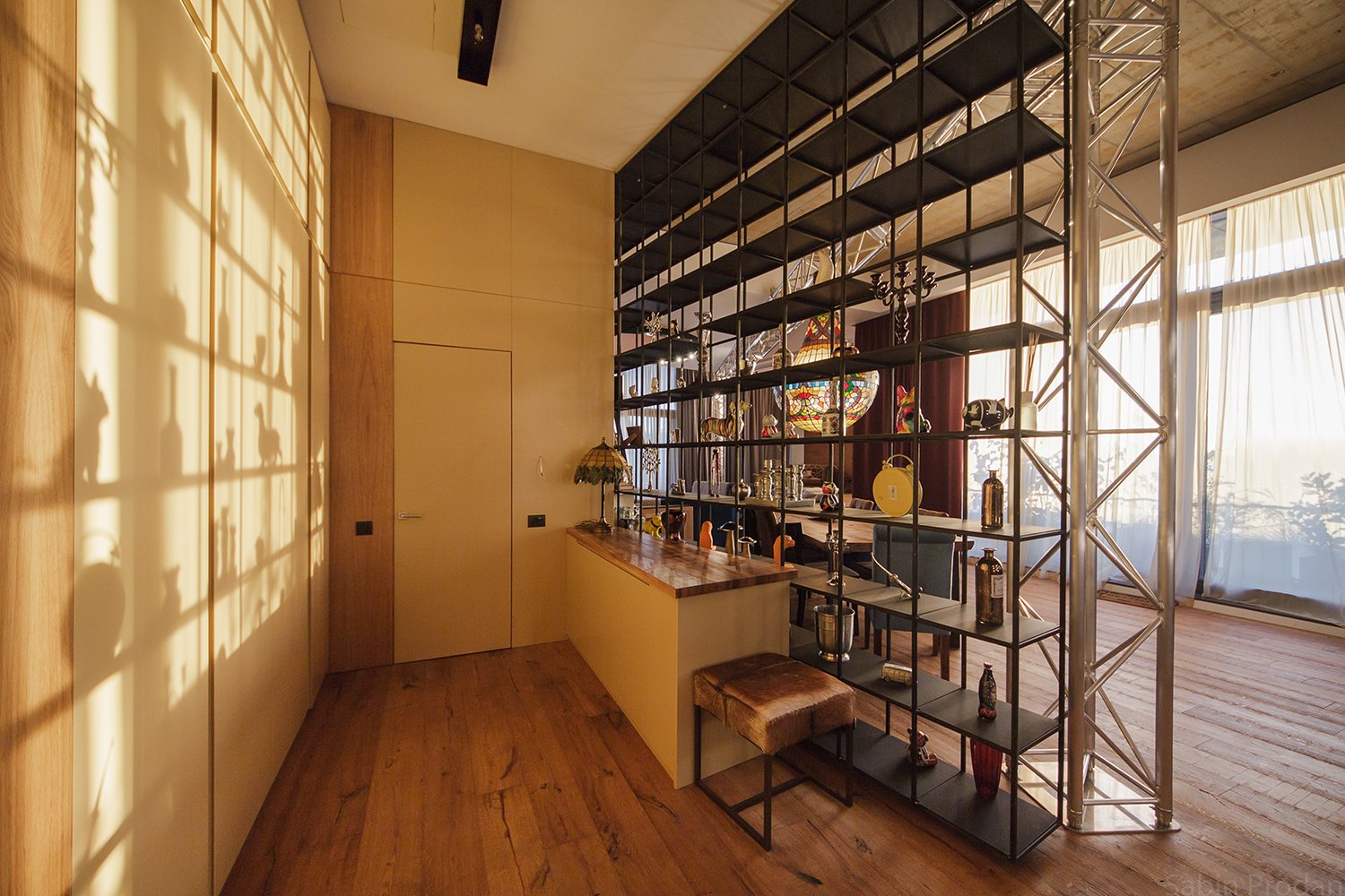 Interior view of a room with wooden flooring, beige walls, and a large metal shelving unit filled with various decorative items, with sunlight streaming through sheer curtains.