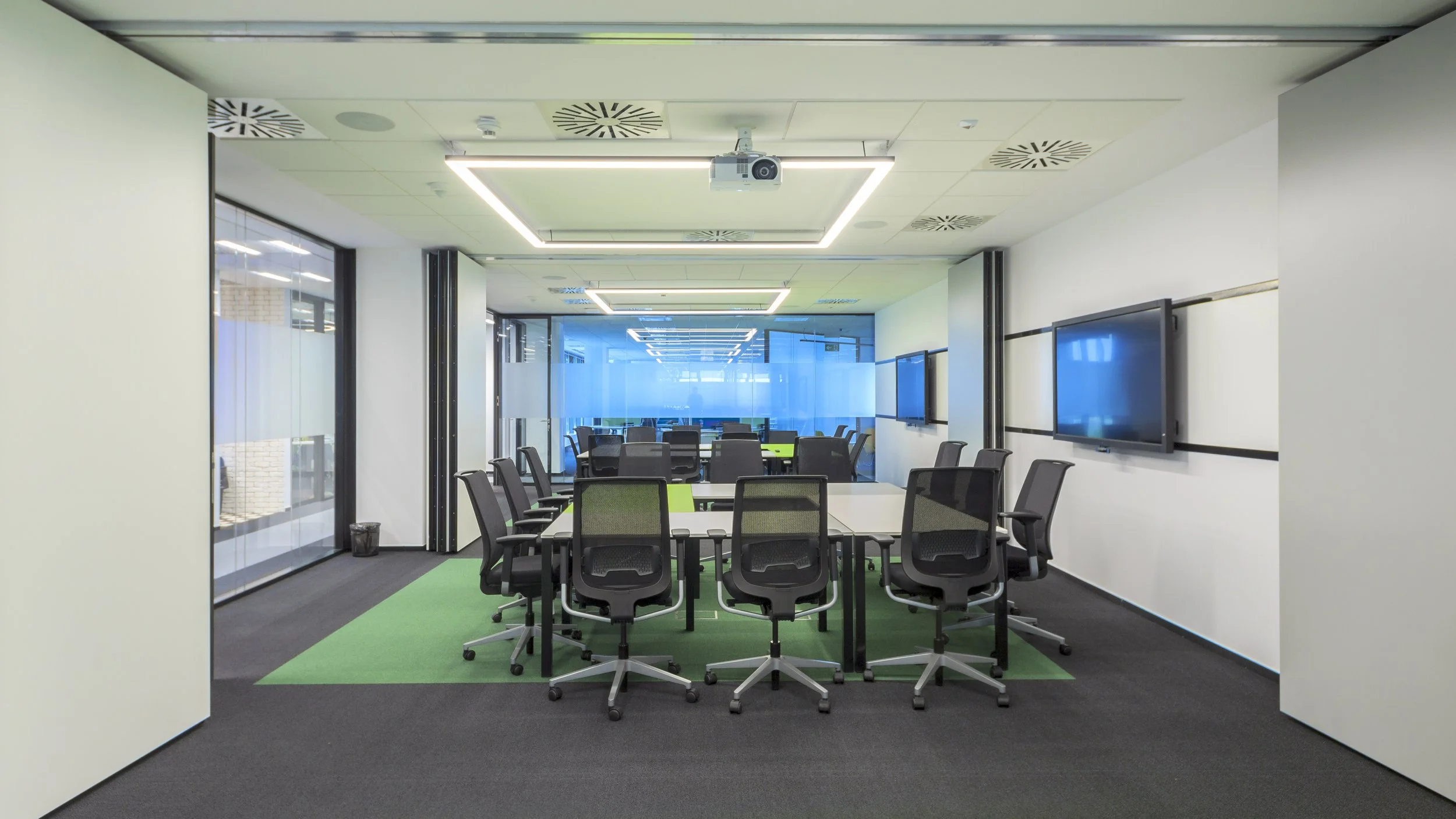 Modern conference room with a long table, black office chairs, wall-mounted monitors, and a ceiling-mounted projector, with a glass wall in the background.