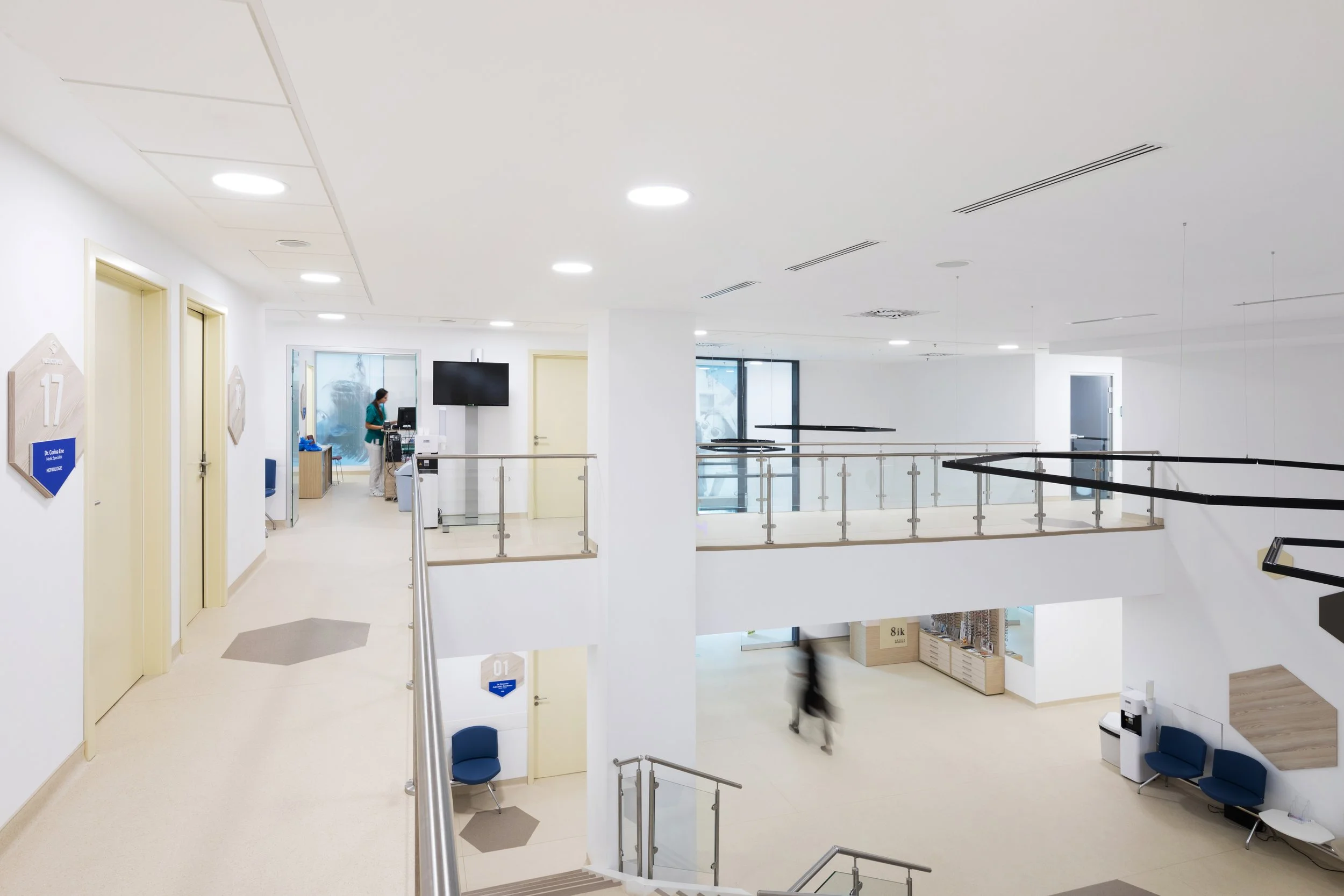 Empty medical facility with white walls, two cream-colored elevator doors, blue seating, a reception area, and a person walking in the background.