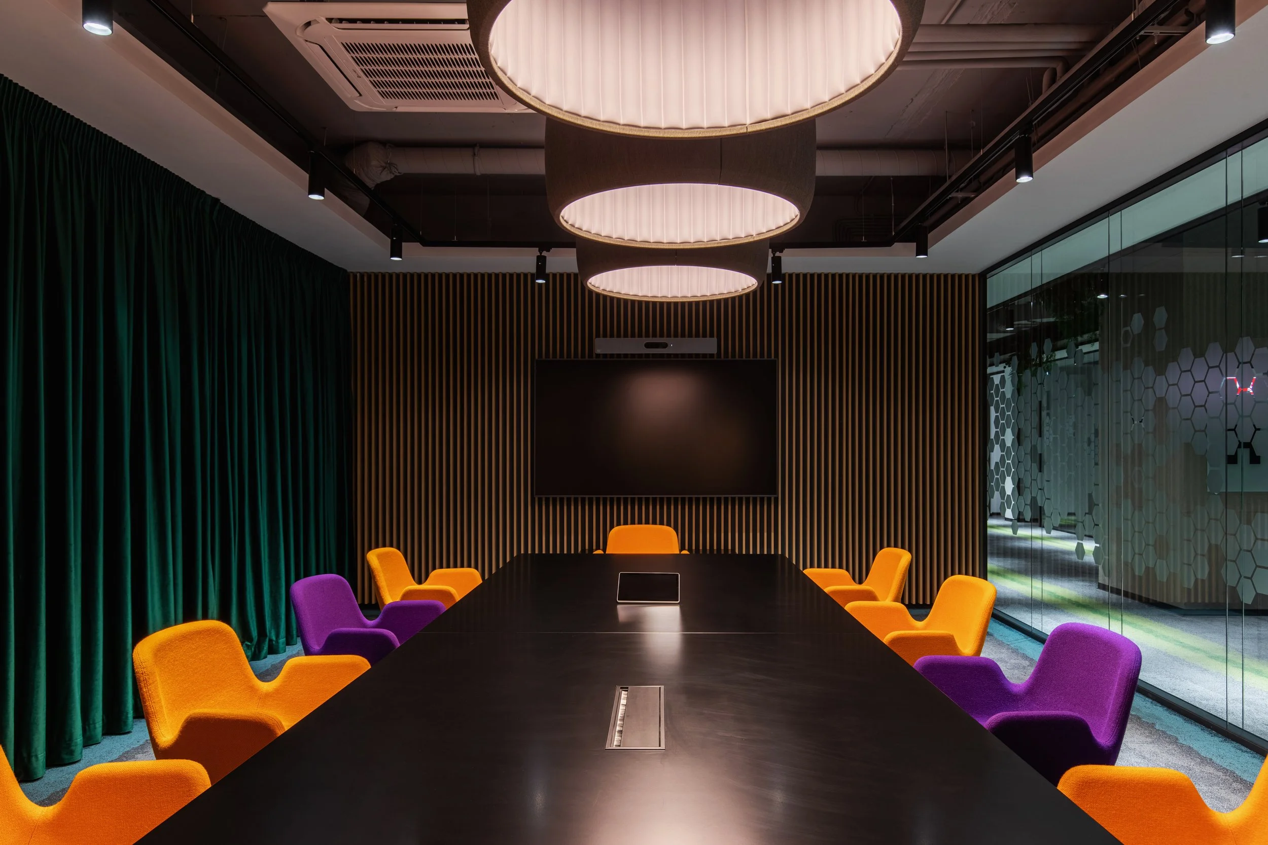 Modern conference room with a long black table, colorful orange and purple chairs, a large flat-screen TV on a wood-paneled wall, and ceiling lighting fixtures.