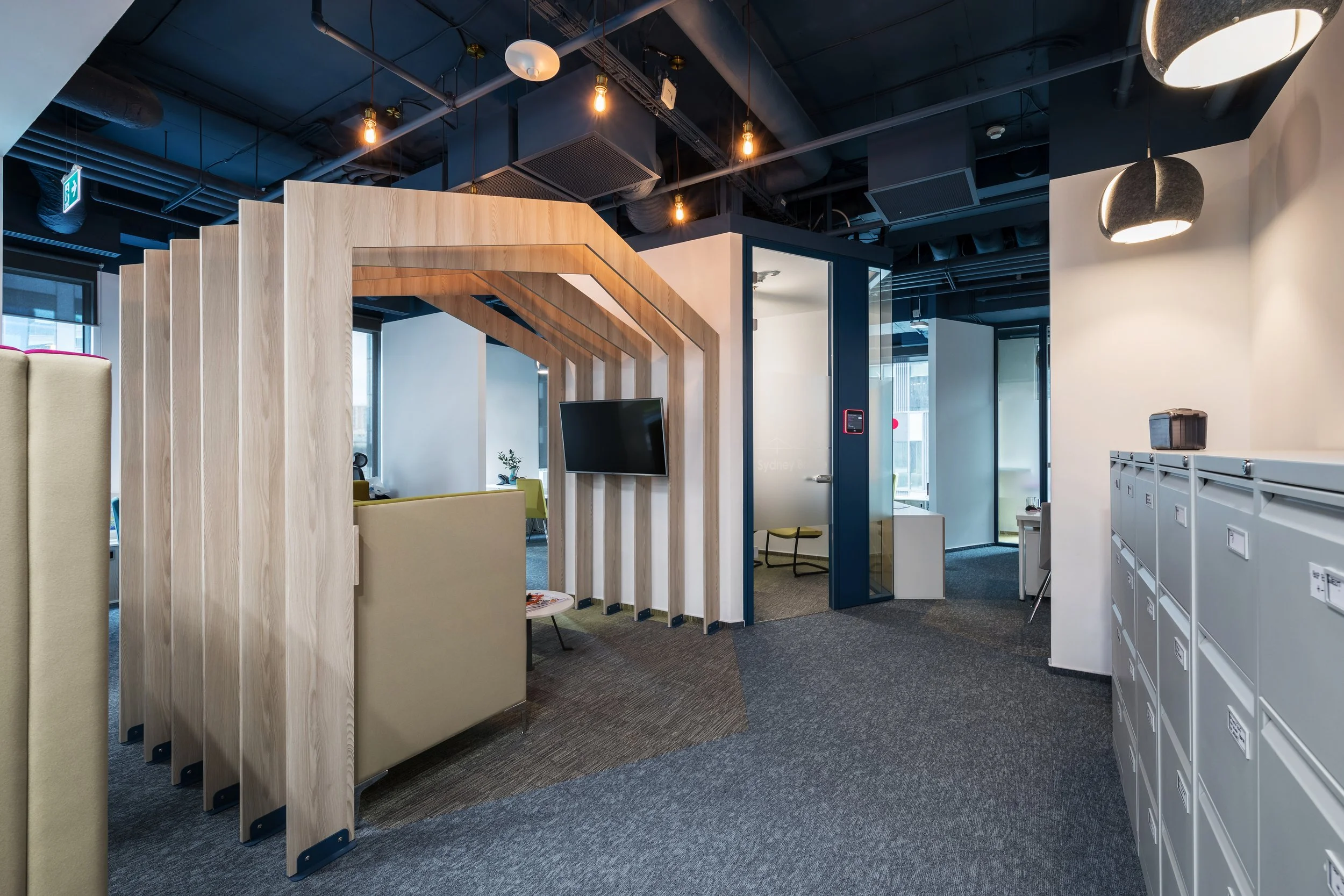 Modern office space with a wooden partition, a wall-mounted TV, lockers on the right, and glass conference rooms in the background.