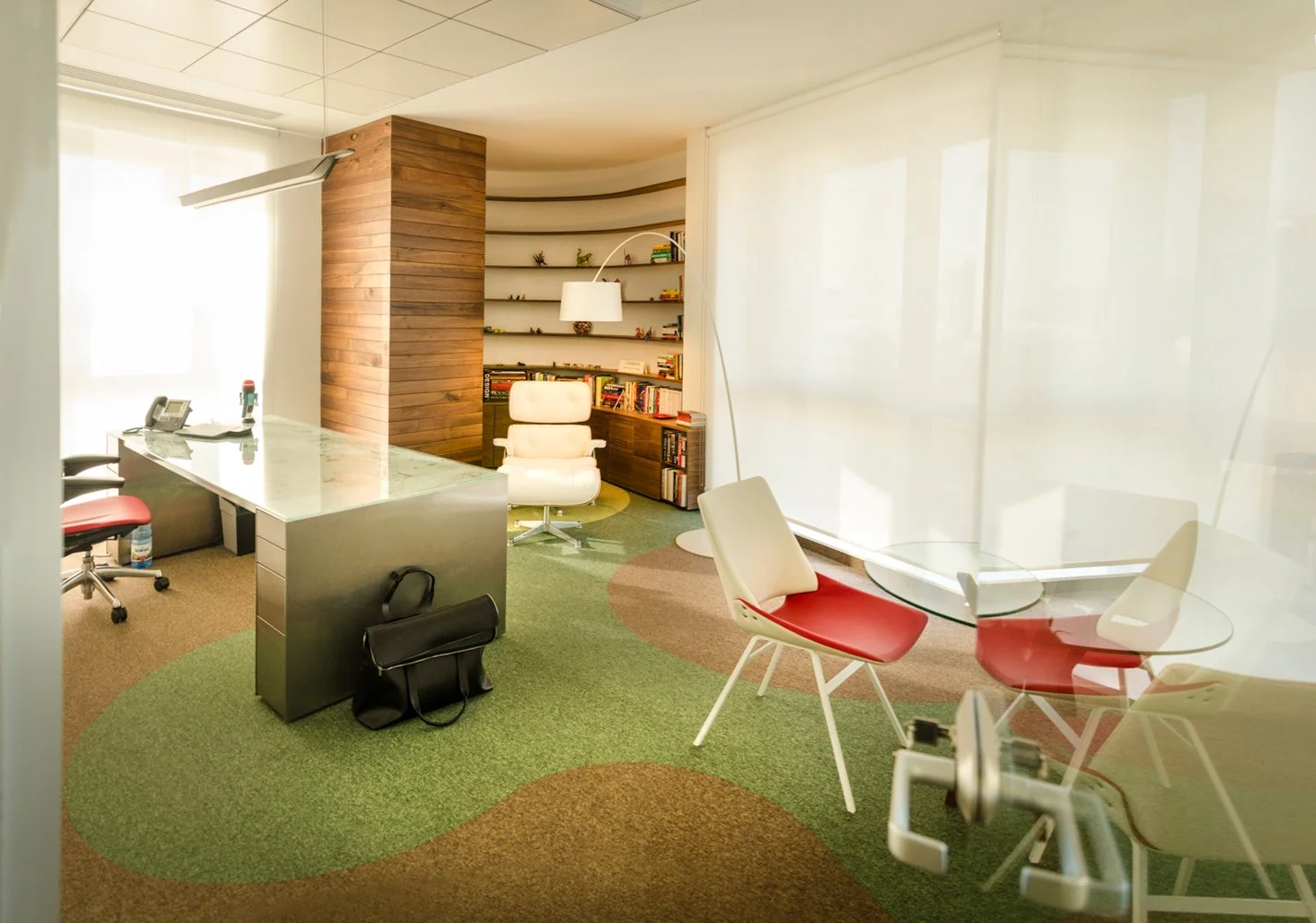 Modern office with a white desk, red and white chairs, a white office chair near a bookshelf, and a glass table with chairs, in a room with green and brown carpet and large windows covered with white blinds.