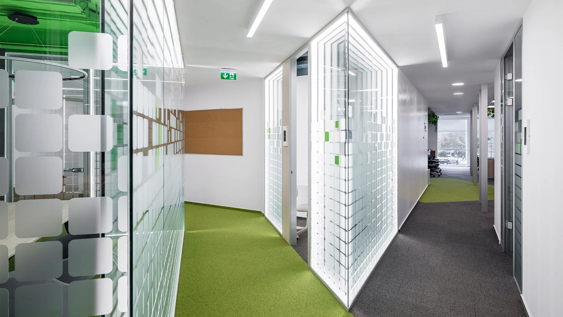 Modern office hallway with glass walls, green and gray carpets, and white ceiling with long strip lights, leading to a room with large windows and an outdoor view.