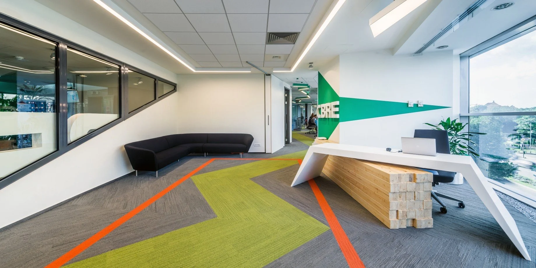 Modern office reception area with a black curved sofa, a white L-shaped reception desk with wooden accents, green and gray geometric carpet, large window with trees outside, and a green wall with white letters.