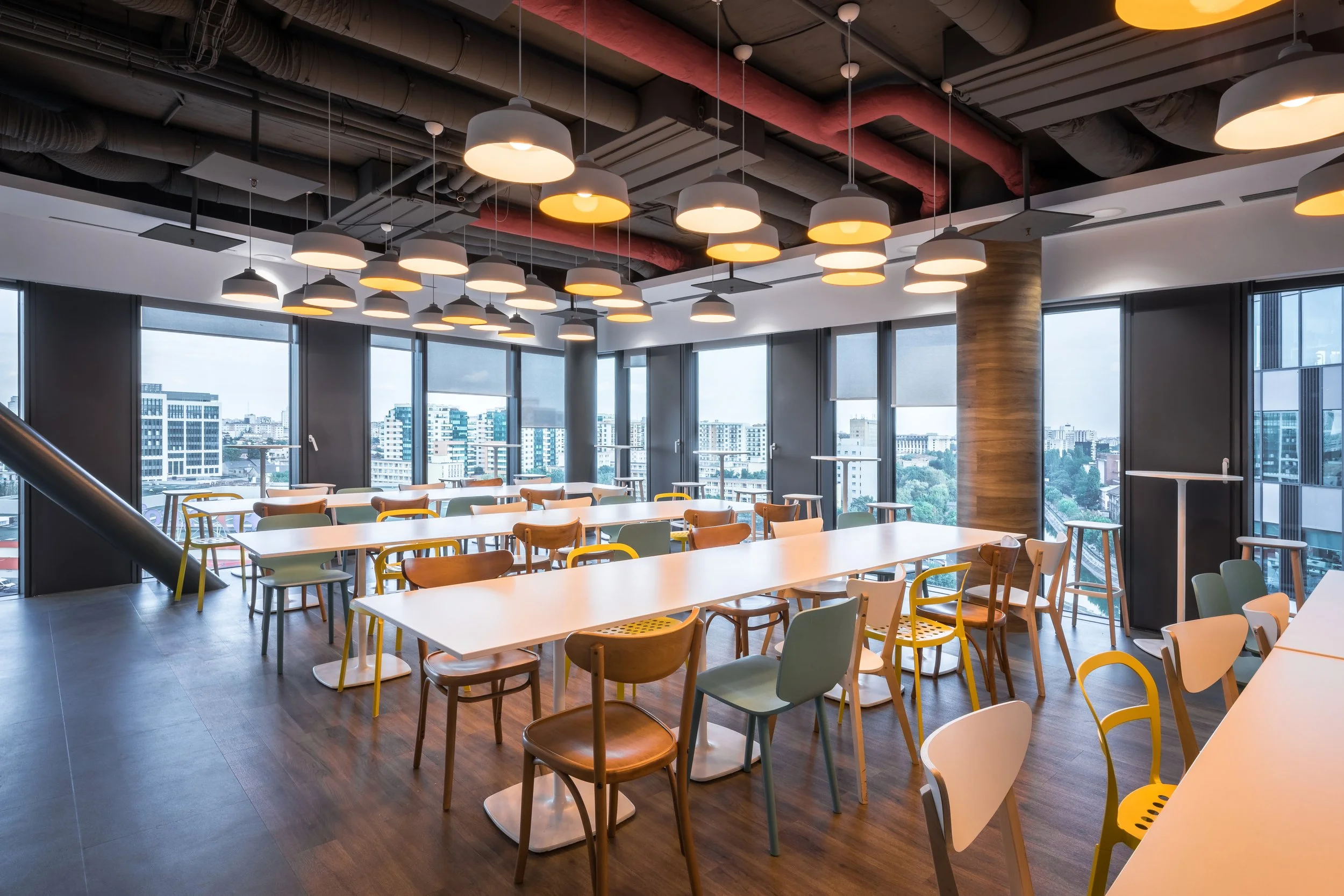 Modern office space with long white tables, colorful chairs, and large windows showing city buildings outside, ceiling with multiple hanging pendant lights.