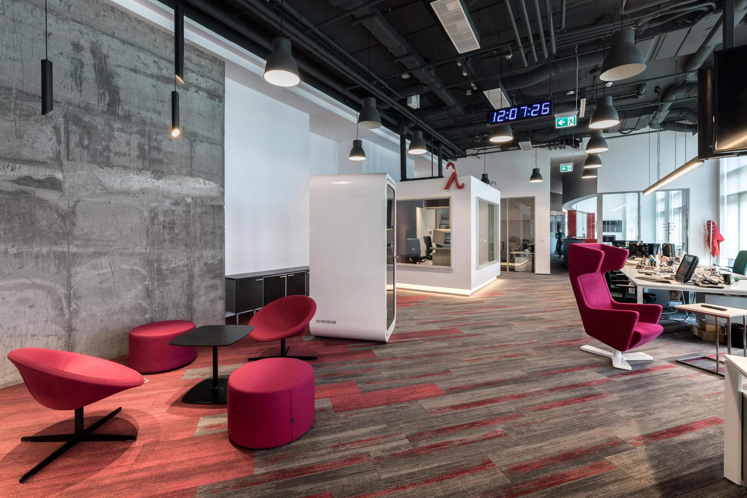Modern office lounge area with pink chairs, a white pod-like enclosure, a large open workspace with multiple desks and computers, and a digital clock on the ceiling showing 12:07:26. The office has an industrial design with exposed ceilings, hanging lights, and large windows.