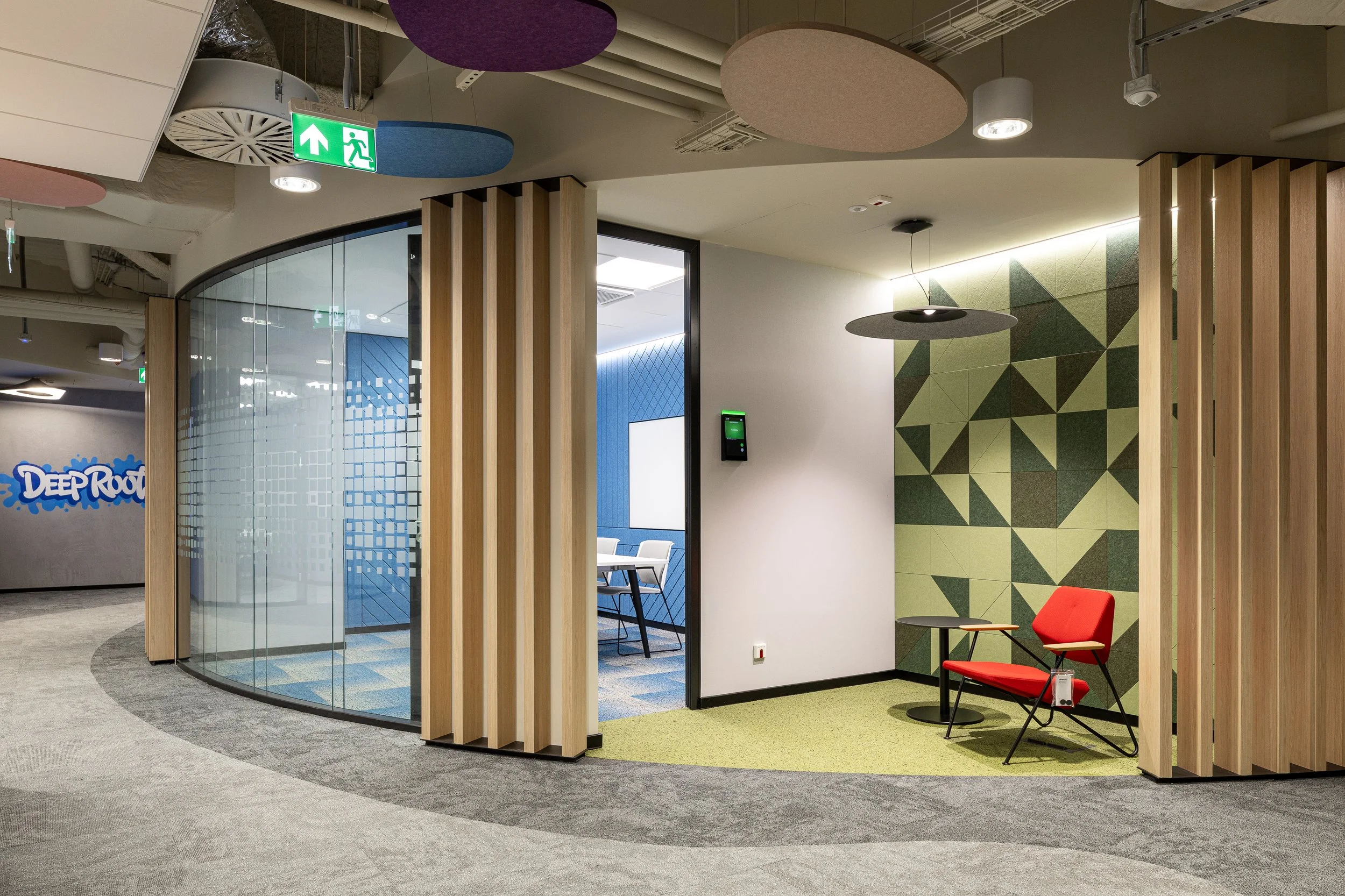Modern office lounge area with red chair, small table, and geometric wall design, separated by vertical wooden slats, with a door leading to a conference room and colorful ceiling panels.