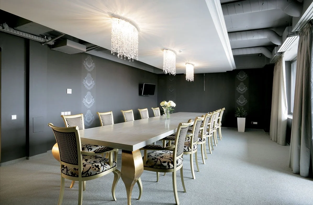 A modern conference room with a long, white table and multiple ornate chairs, floral centerpiece, gray walls, large window with curtains, and three hanging chandeliers.