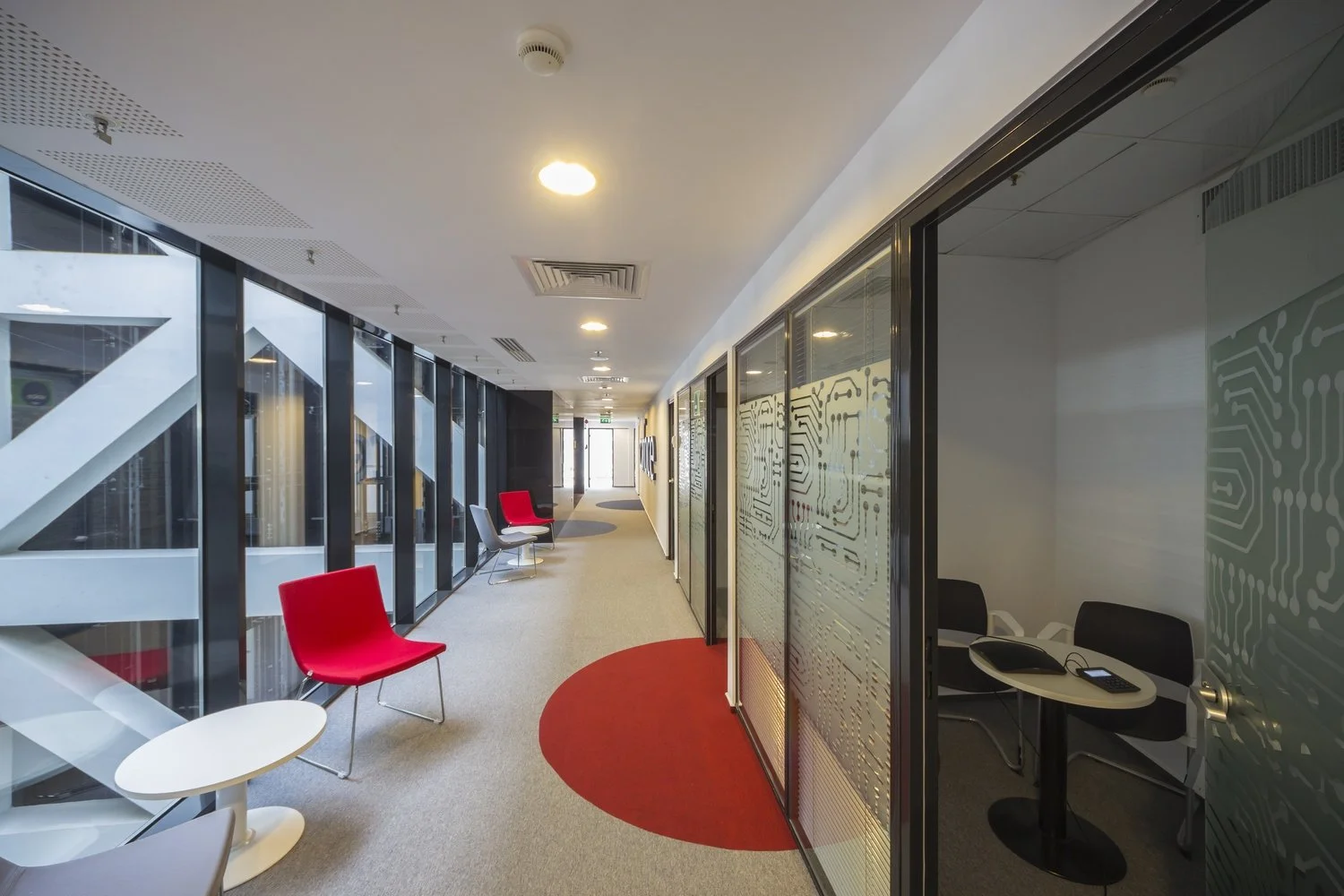 Modern office corridor with glass walls, red and gray chairs, small white tables, and a curved red rug.