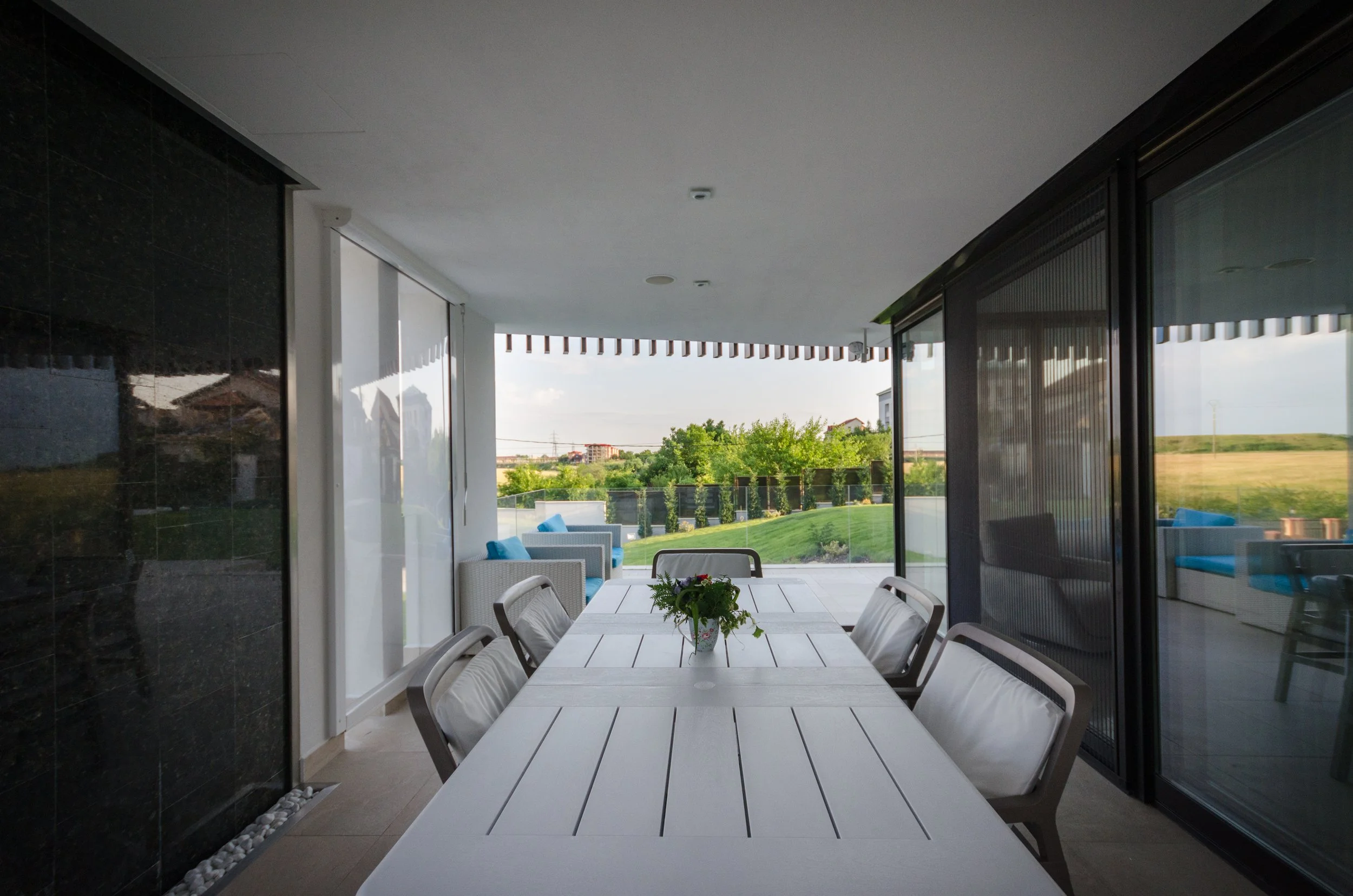 Outdoor patio with a white dining table, six chairs, a small potted plant in the center, and a seating area with white wicker chairs and blue cushions, overlooking greenery and distant buildings.